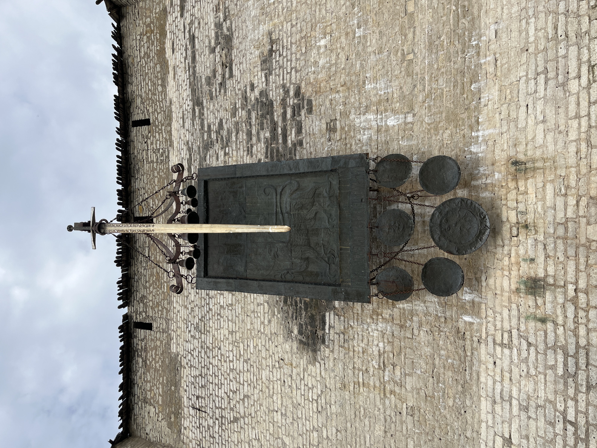 A medieval sword monument is affixed to a stone wall in Pskov, Russia. The monument features a long, narrow sword pointing downwards, suspended by a decorative metal frame holding a set of bells. Below the bells hangs a large, dark rectangular plaque with intricate relief carvings. Dangling from chains below the plaque are several round, metal objects, possibly shields or gongs. The stone wall is constructed of roughly cut, light-colored bricks, with some signs of weathering and staining. The top of the wall is topped with wooden planks, some of which appear damaged. The sky above is overcast.