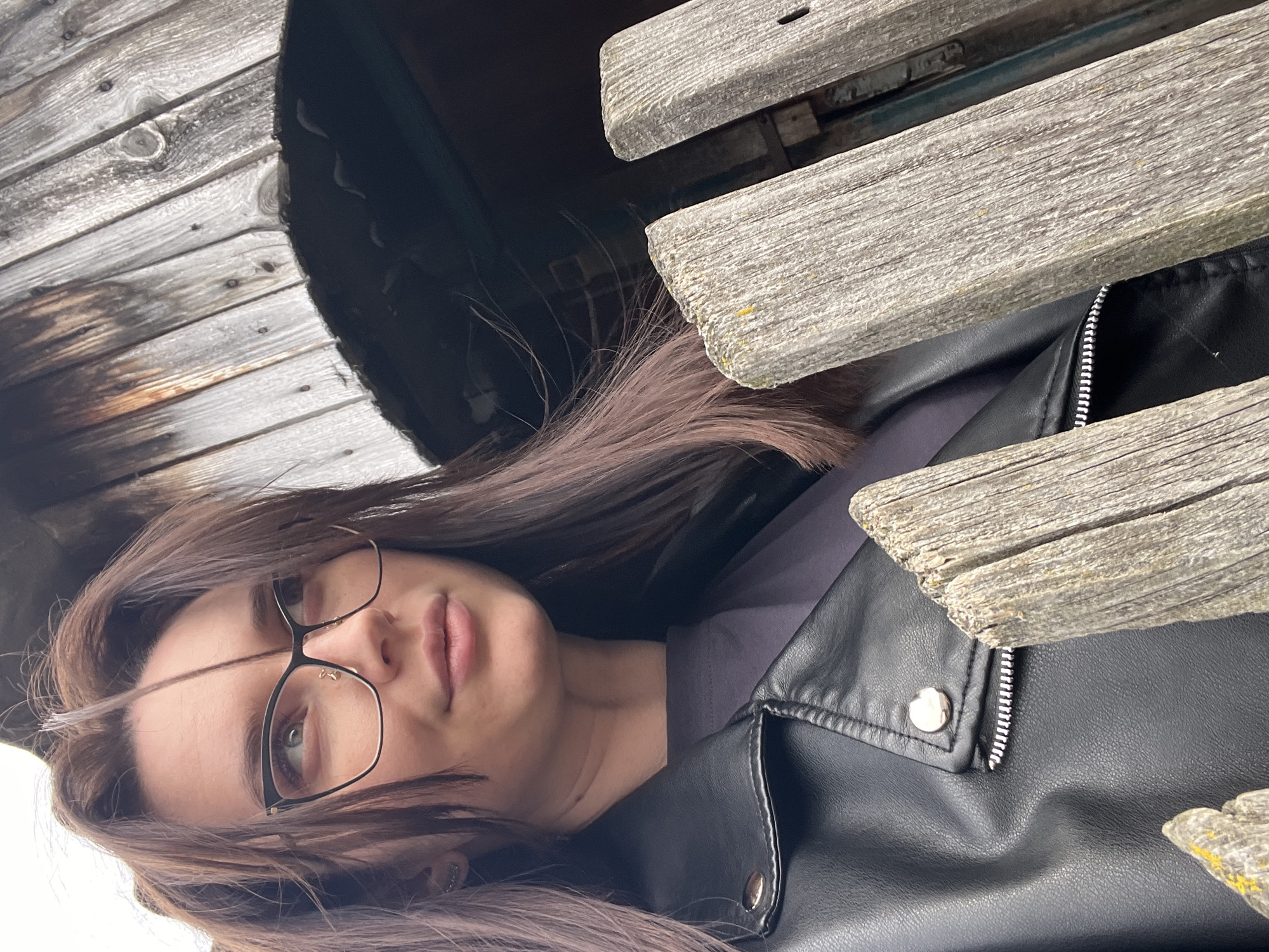 A person wearing glasses and a black leather jacket is positioned underneath what appears to be a wooden structure, with light-colored wooden planks or boards visible above and to the left. The background is difficult to discern but seems to include weathered wooden surfaces. The person is looking upwards with a neutral expression. The image seems to be taken at an unusual angle. It is not possible to confirm this photo was taken in Usol'tseva, Russia.