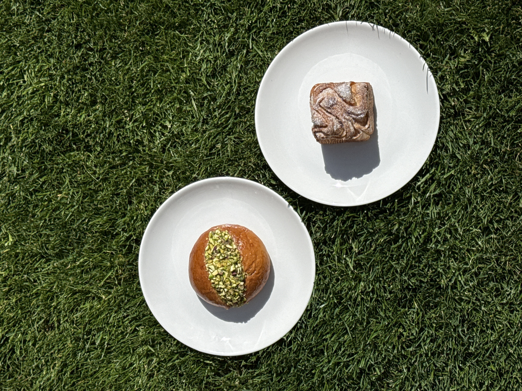The image shows two white plates sitting on a lawn. On the top right plate is a square-shaped pastry. On the bottom left plate is a round pastry topped with chopped green nuts, possibly pistachios. The background is green grass. The location is unknown, but the description does not indicate the city of Belgrade, Serbia.