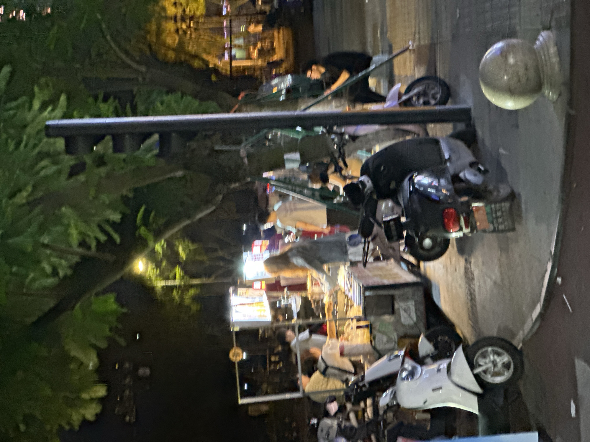 This nighttime image depicts an urban street scene in Chongqing High-tech Zone, China, characterized by active street vending, pedestrian presence, and parked motorbikes under artificial illumination.
The environment is dimly lit by streetlights and bright vendor stall lighting, casting shadows and highlighting the immediate foreground and middle ground. Large trees with dense green foliage are prominent throughout the scene, obscuring parts of the sky and background structures. The ground consists of concrete or asphalt pavement with visible curbs and a spherical stone bollard in the lower right foreground.
Approximately six to eight individuals are present. In the mid-ground, a group of people is gathered around two brightly lit mobile vendor stalls. One person with long, light-colored hair, wearing a light-colored jacket, is seen reaching towards a stall. Other individuals, including one in a light grey shirt, are positioned near or behind the stalls, suggesting vendor or customer roles. To the far right, a male individual with dark hair and glasses, wearing a dark shirt and pants, is seated on a low structure, leaning against a metal support pole near a tree. His gaze appears directed towards the center of the scene.
Multiple two-wheeled vehicles are parked on the pavement. A white scooter with silver wheels is prominently positioned in the lower left foreground, partially obscuring other vehicles. Further right, a dark-colored scooter with a red taillight and a partially visible license plate is parked near the spherical bollard. Other scooters are visible in the mid-ground, interspersed among the people and vendor stalls. A dark, vertical pole, possibly a traffic light or utility pole, stands on the right side of the image, partially surrounded by tree foliage. The vendor stalls themselves are constructed with metal frames, displaying various items illuminated by internal lights. A round sign is visible above the left-most vendor stall.