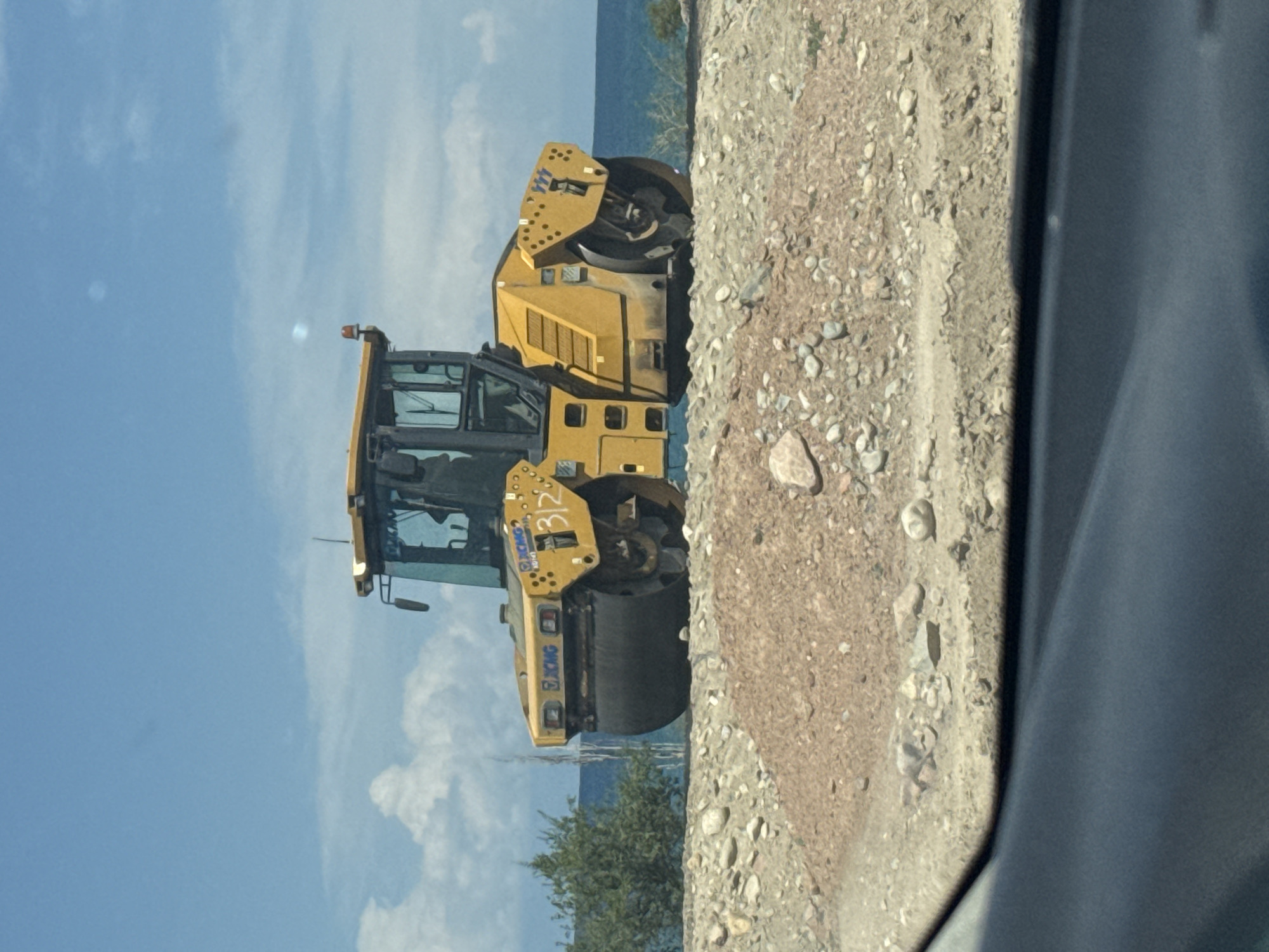 A yellow road roller is parked on a gravel embankment. The road roller has "XCMG" branding and the number "312" painted on its side. In the background, a body of water is visible, with trees and bushes on the left and right sides of the embankment. The sky is blue with scattered white clouds. The foreground shows a dark, blurred surface, possibly the interior of a vehicle. The image appears to have been taken outdoors in a location that could be Tong, Kyrgyzstan, given the context, characterized by a sunny day with a clear blue sky and a mountainous or hilly landscape.