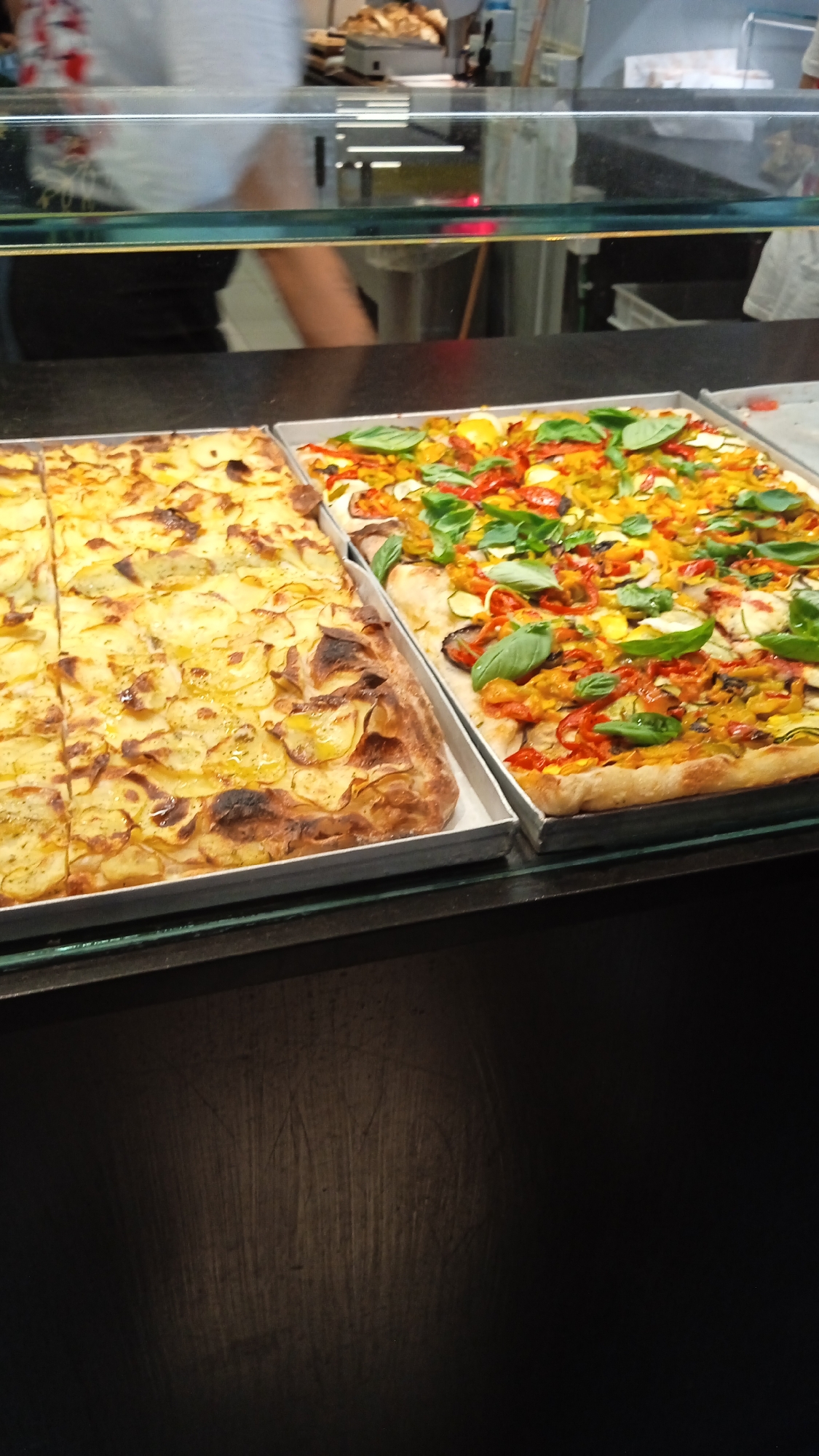 This is a close-up, eye-level, slightly angled shot of two large rectangular pizzas displayed in metal trays behind a glass counter. The pizza on the left is topped with thinly sliced potatoes and appears to be cooked to a golden brown with some darker, crispier edges. The pizza on the right is a vegetable pizza with a colorful assortment of toppings including red bell peppers, yellow bell peppers, zucchini, and fresh basil leaves. Both pizzas are cut into rectangular portions. The background is blurred, showing a commercial kitchen or food service environment with a glass barrier and some indistinct figures. The lighting is bright, illuminating the food. The setting is likely a bakery or pizzeria in Milan, Italy.