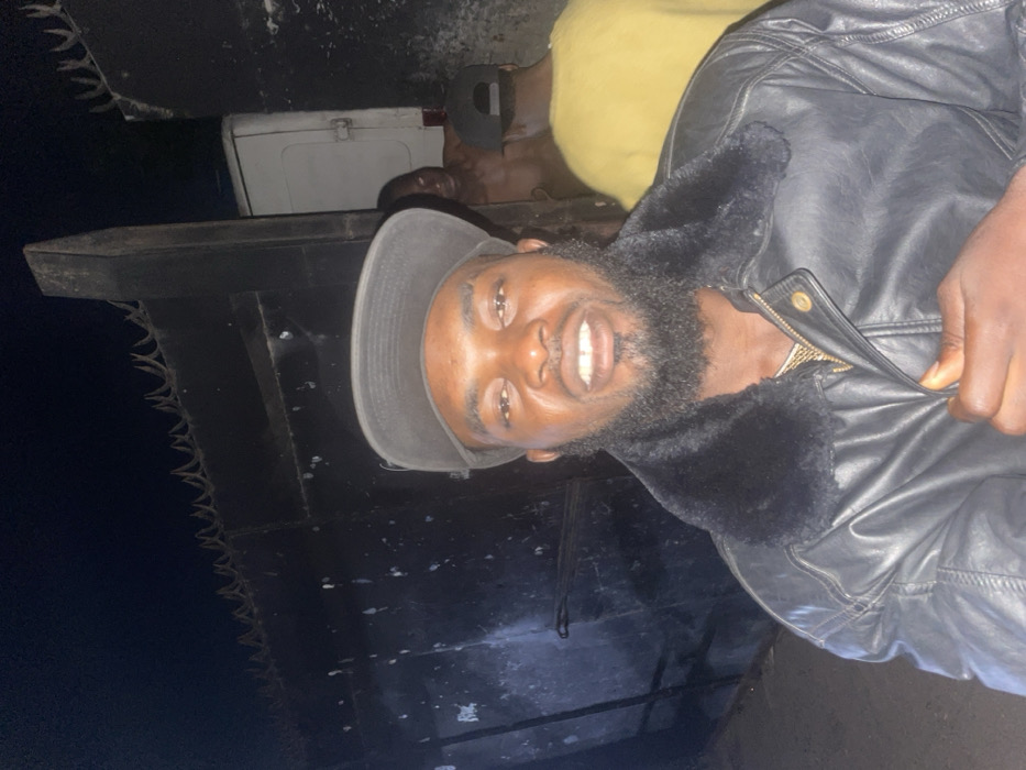 A smiling man wearing a black leather jacket with a fur collar and a gray cap is partially visible. The background features a dark, textured surface, possibly a ship's hull with protruding spikes. A white box and a person in a yellow shirt are also partially visible behind the man. The image is rotated.