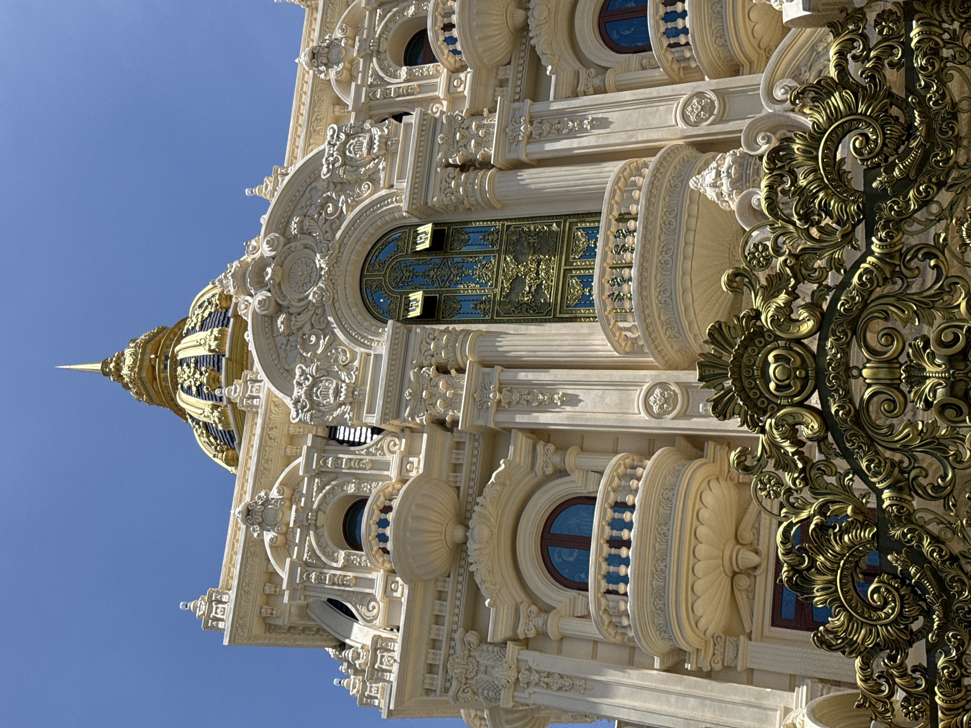 A low-angle, vertically-oriented shot presents the intricate facade of a building, set against a clear blue sky. The building is notable for its ornate architectural design, including decorative columns, arched windows, and a golden dome at the top left of the frame. The facade is predominantly light in color, possibly white or a pale cream, and exhibits elaborate carvings and moldings. The windows are framed in various designs, some with blue-tinted glass. A decorative metalwork structure, possibly a gate or railing, is visible at the right edge, featuring a bronze-like finish and complex floral patterns. The architectural style and ornamentation suggest the building is a temple or a palace located in Da Lat, Vietnam.