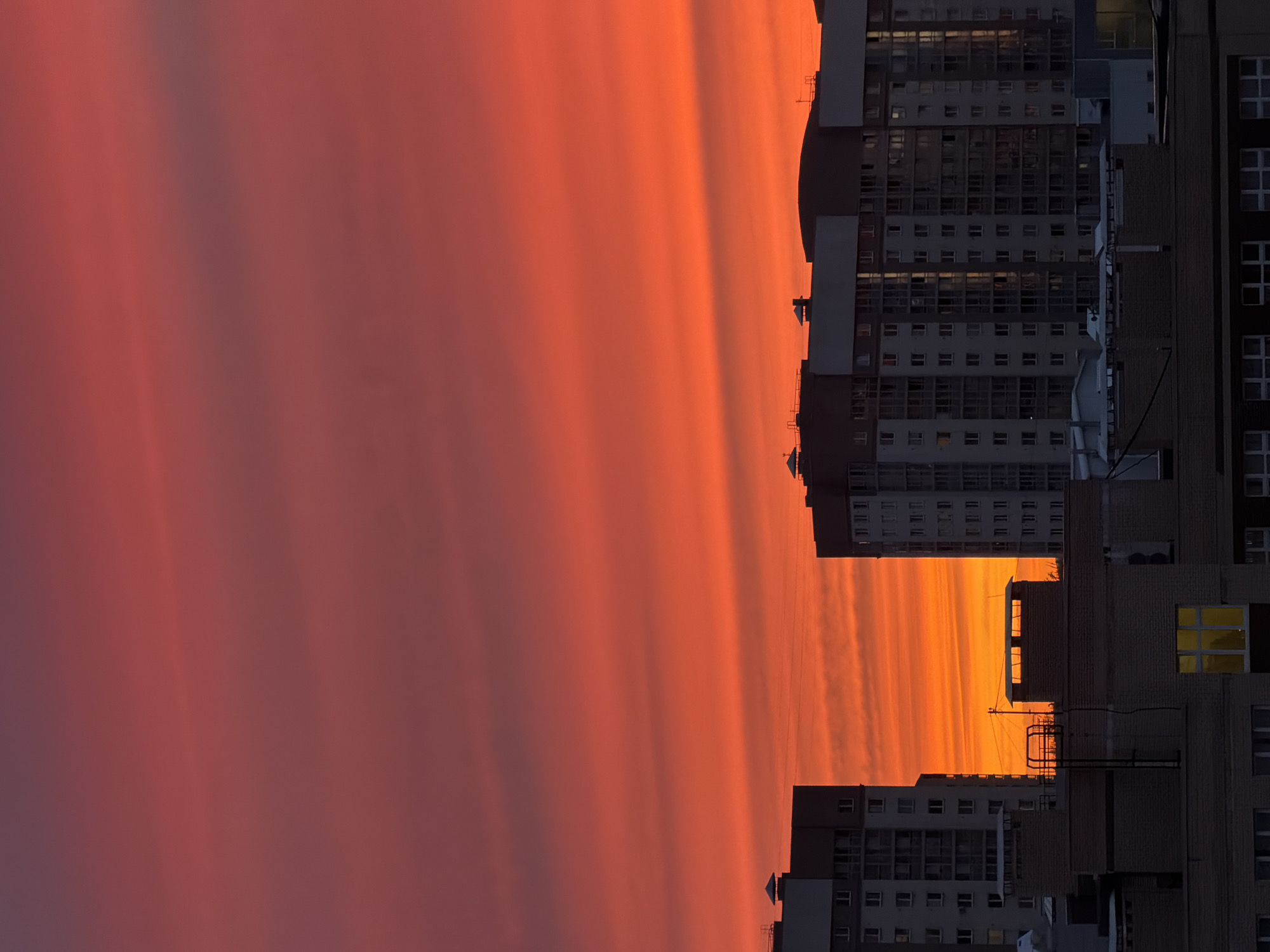 The image depicts a vibrant sunset over a cityscape in Saint Petersburg, Russia. The sky is ablaze with shades of red, orange, and purple. Silhouetted buildings with multiple stories are visible in the foreground. Some windows are illuminated, indicating activity within. The perspective is unusual, possibly a view from a tall building, showing the city at dusk.