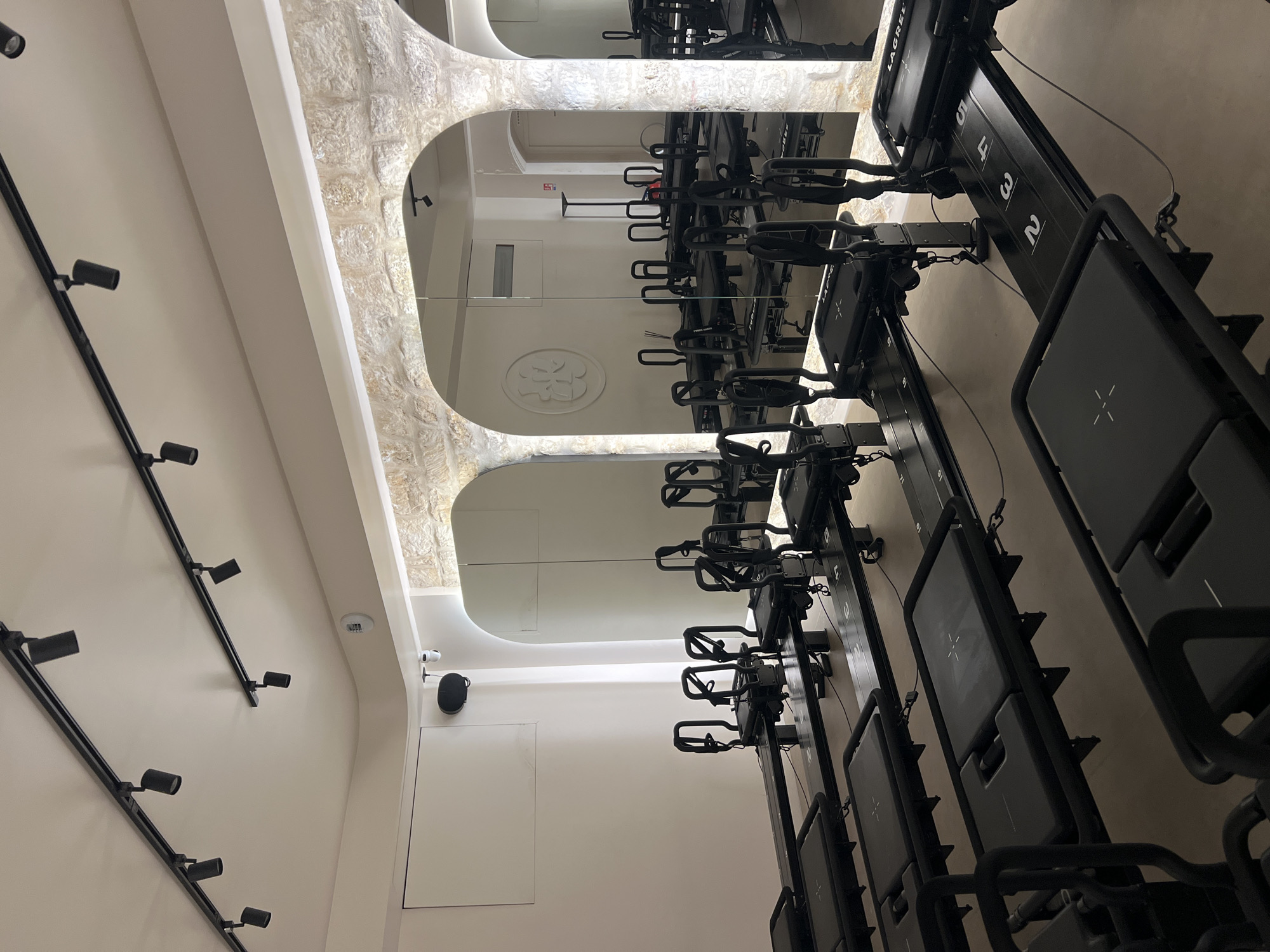 The image shows the interior of a Lagree Fitness studio, likely located in Paris, France. The studio is characterized by exposed stone arches along one wall, which are mirrored on the opposite side, creating a sense of symmetry and spaciousness. Rows of black Lagree Fitness machines are arranged in the center of the room, positioned perpendicularly to the walls. The machines are numbered and appear to be ready for use. The color palette is dominated by neutral tones, with white walls and ceilings contrasting against the black equipment. A track lighting system runs along the ceiling, illuminating the space. A minimalist design with white walls, white ceiling, and black equipment, emphasizing functionality and modern aesthetics.