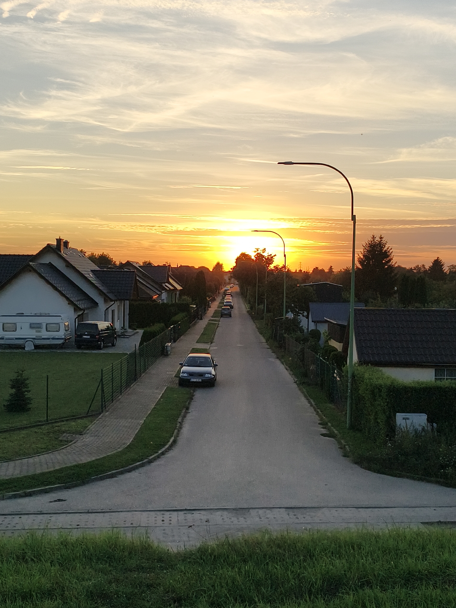 The image depicts a street scene at sunset in Tczew, Poland. A residential street, lined with houses and trees, stretches into the distance. The sky is ablaze with the warm colors of sunset, with hues of orange, yellow, and pink painting the clouds. Several cars are parked along the street, and a camper van is visible in the driveway of one of the houses. The street is paved, with sidewalks on either side. Lampposts are spaced along the street, and a fence runs alongside the sidewalk. The overall impression is one of a quiet, peaceful evening in a suburban neighborhood.