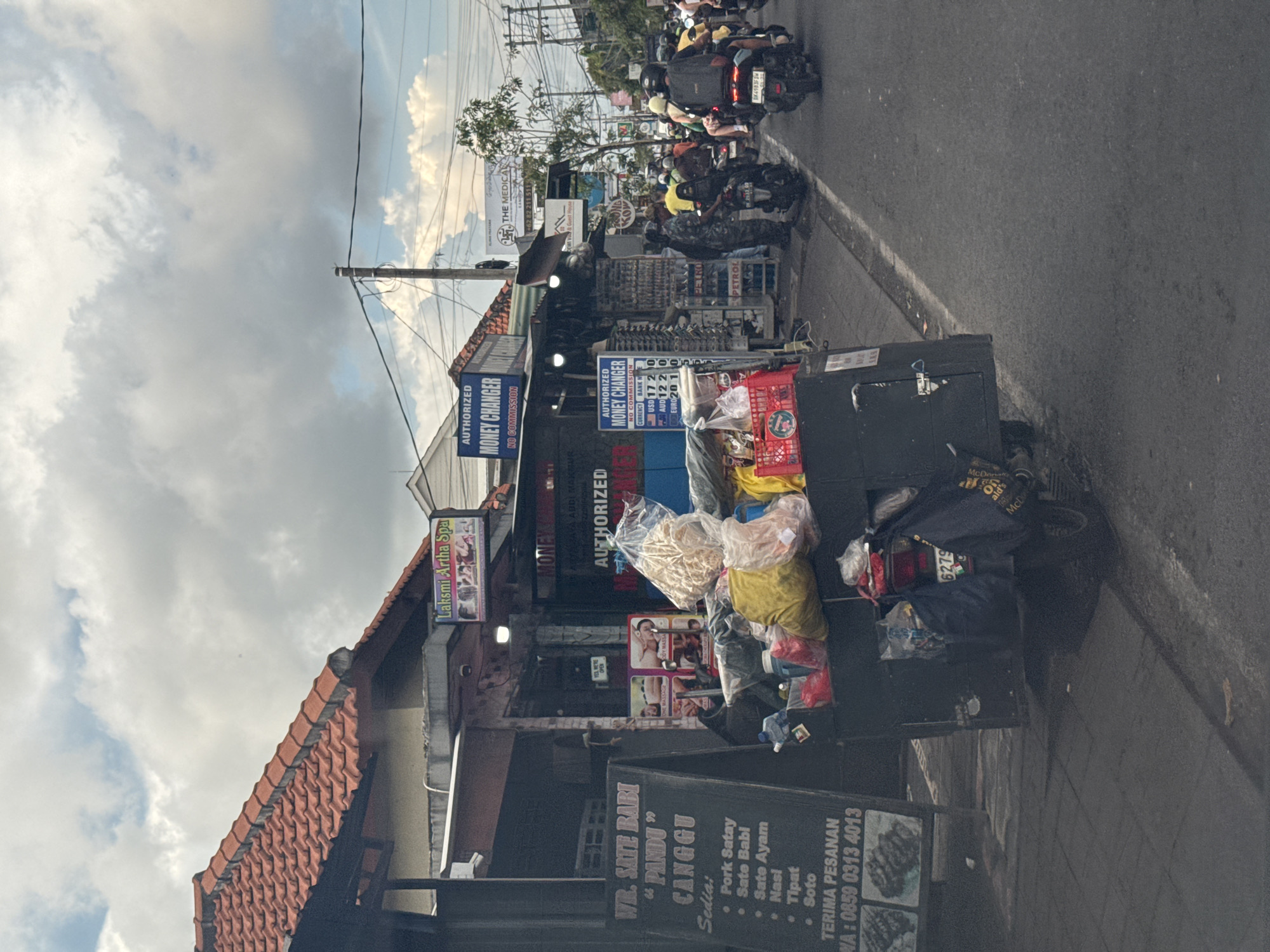 The image depicts a bustling street scene in North Kuta, Indonesia, captured during the daytime under an overcast sky. The perspective is from the roadside, looking across an asphalt road towards a row of shops and businesses.
On the right side of the road, a line of motorbikes, many with riders wearing helmets, suggests a busy traffic flow. One motorbike clearly shows a license plate number, "DK4983F SW." The road features a visible white dividing line.
The left side of the frame is dominated by various street-side establishments. In the foreground, a food stall or small restaurant is visible, identified by a large chalkboard sign that reads "WR. SATE BABI 'PANDU' CANGGU." The menu lists items such as "Pork Satay," "Sate Babi," "Sate Ayam," "Nasi," "Tipat," and "Soto." It also provides contact information for orders: "TERIMA PESANAN WA: 0859 0313 4013." Nearby, a utility cart holds various items in plastic bags, and a motorbike with "McDonal's" text on a bag and license plate "6279" is parked.
Further along the street, multiple signs advertise "AUTHORIZED MONEY CHANGER NO COMMISSION," with one displaying currency exchange rates: "USD 172," "AUD 122," and "EURO 201." Other visible business signs include "Laksmi Artha Spa," "THE MEDICAL," and a partial sign for "ABDI MAK." Power lines are strung across the street above the shops, and the buildings feature traditional red-tiled roofs. The scene conveys the lively commercial activity typical of a street in this region.