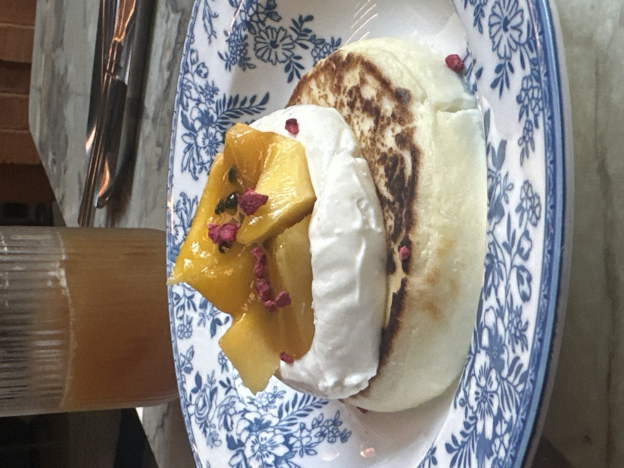 A close-up shows a dessert dish presented on a blue and white patterned plate. The dessert consists of a pancake-like base topped with whipped cream and diced mango, garnished with small, dark-colored seeds and flecks of what appears to be crushed dried flowers or fruit. A glass of orange-colored liquid sits adjacent to the plate, suggesting it might be a beverage intended to accompany the dessert. The style of plating and the decor hints at a café setting, but the specific location within Novosibirsk, Russia, cannot be determined from this image alone.