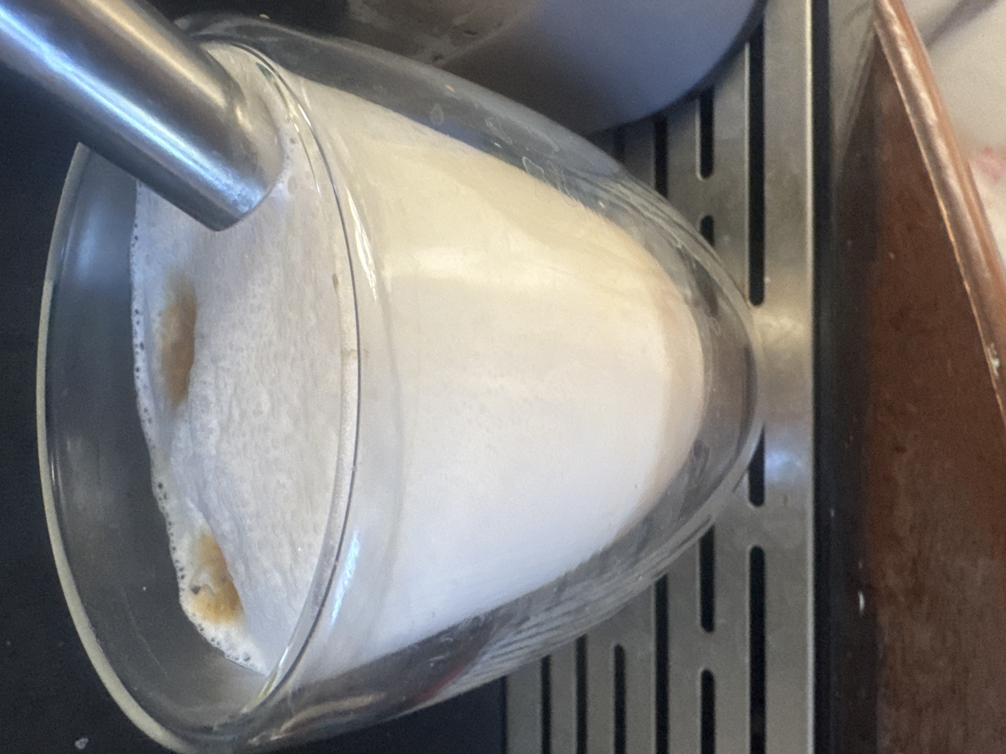 In Sochi, Russia, a close-up shot captures a glass of frothed milk being prepared for a beverage. The glass, made of clear, double-walled glass, is filled with a thick layer of white foam. A metallic wand, likely from an espresso machine, is partially submerged in the foam, suggesting the frothing process is either in progress or has just concluded. A small amount of dark liquid, possibly coffee, is visible on the surface of the foam, indicating that this is likely the initial stage of making a coffee drink like a latte or cappuccino. The glass sits on a metallic grate, typical of coffee-making equipment. The background is dark and indistinct, focusing all attention on the beverage.