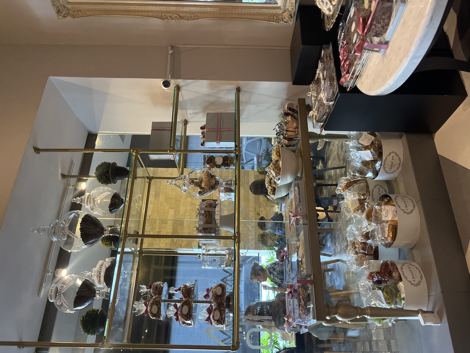 This indoor scene, seemingly taken within a shop or boutique in Puebla, Mexico, features a glass and gold metal shelving unit filled with various packaged treats. The name "Chocol'art" is visible on many of the containers and packaging. Glass jars filled with dark-colored items, potentially chocolates or other confections, sit on the upper shelves, along with small potted plants. The middle and lower shelves hold individually packaged items adorned with ribbons. Reflections in the glass shelves reveal glimpses of people browsing the shop, including a woman in a floral shirt and another individual with longer hair. Above the shelving unit, a framed piece of art is partially visible at the top of the frame, mounted on the wall. A security camera is also visible near the top of the frame. The arrangement suggests a retail setting specializing in gourmet or artisanal sweets, likely located in the city of Puebla, Mexico.