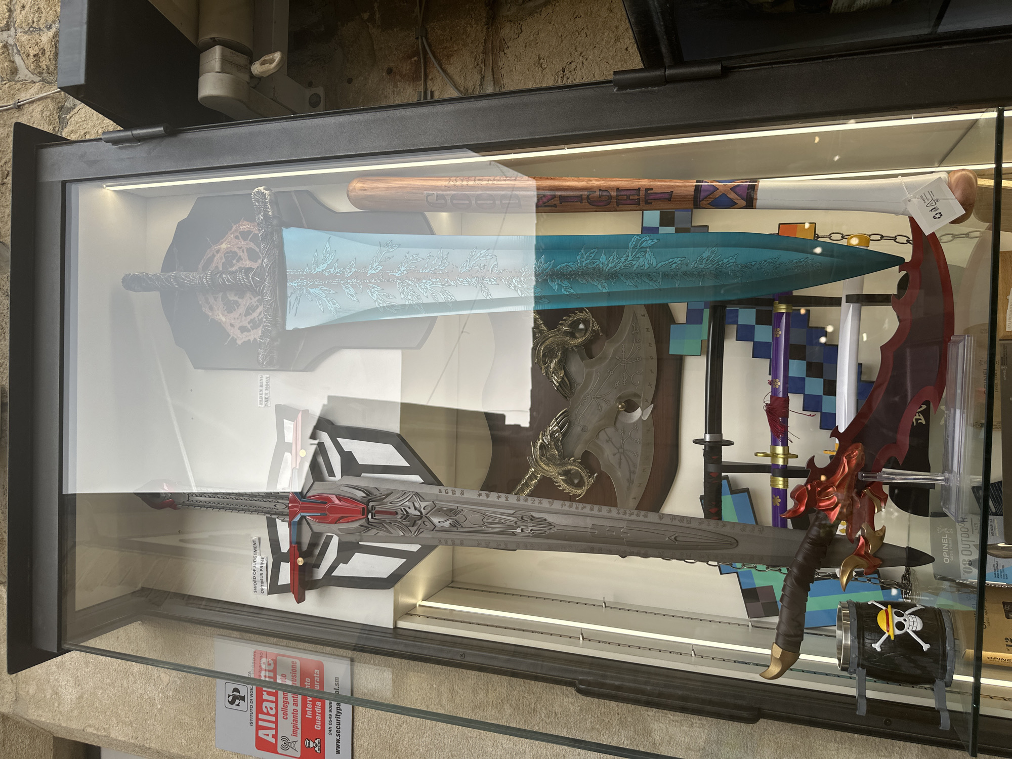 The image shows a display case in a shop in the city of San Marino, San Marino. The display case contains various replica weapons, including a large sword with a red and gray hilt and blade, a large blue-green sword with decorative etchings, and a wooden baseball bat with text that appears to read "GOOD NIGHT." Behind these items, there is an axe with a dragon motif and a shield-shaped symbol that resembles the Autobot logo from Transformers. Other items visible include smaller swords, a mug with the One Piece Jolly Roger logo, and various labels and tags. The display case is made of glass and metal, and it is illuminated by interior lights. The background consists of a stone wall.