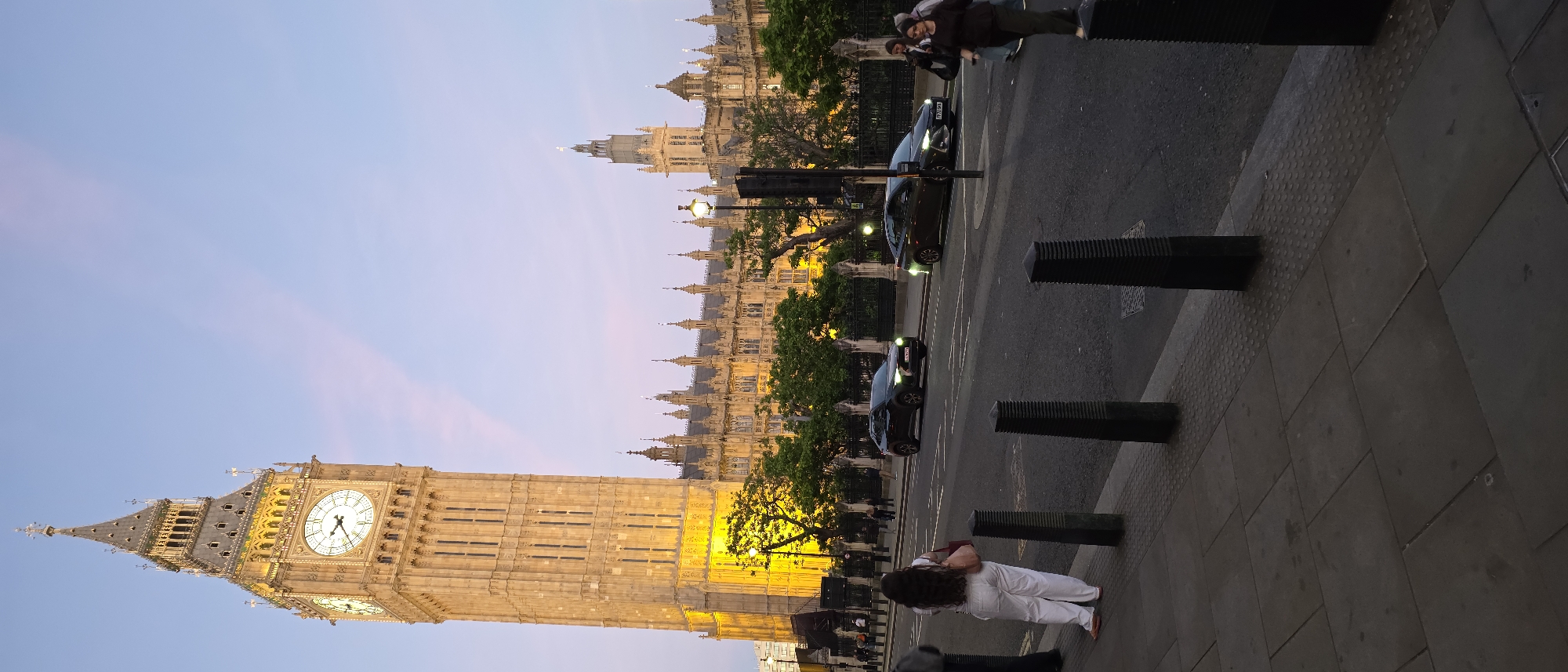 This is a full shot, vertical, outdoor, natural light photograph taken in London, United Kingdom. The primary subject is the Elizabeth Tower, commonly known as Big Ben, which is illuminated by yellow lights from below. The tower is a tall, ornate, stone structure with a clock face on one side. To the right of the tower is the Palace of Westminster, a large Gothic Revival building. In the foreground, there is a sidewalk with patterned paving stones and a row of black bollards. Across the street, there is traffic, including two dark-colored cars, and several pedestrians walking. The sky is a pale blue with streaks of pink, suggesting it is either sunrise or sunset. A woman in white pants and a pink purse is standing on the sidewalk, facing away from the camera.