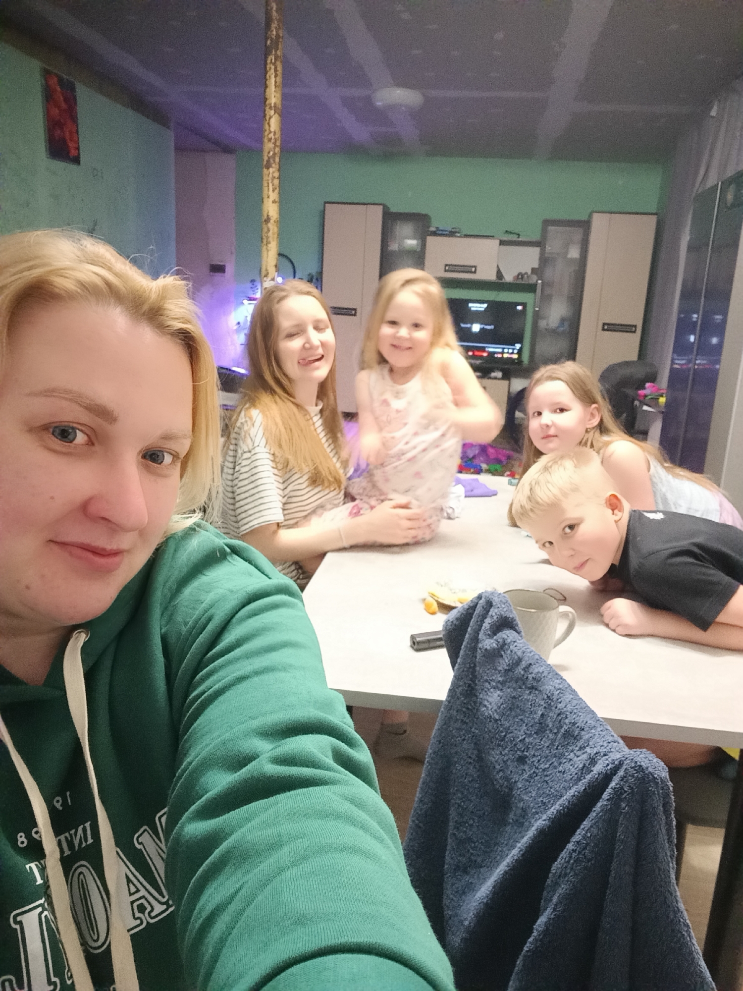 The image shows a woman taking a selfie with four children in a room. The woman is wearing a green hoodie and has blonde hair. Behind her are four children; two girls and one boy are at a table, and one older girl is sitting behind the youngest girl. A blue towel is draped over a chair in the foreground. A table with a cup and a few small objects is visible. In the background is a television set and what appears to be a wall unit or cabinet. The room's walls are painted with a blue-green shade, and there is an exposed ceiling.
It is not possible to confirm the location of this picture with any degree of certainty, as there is a lack of identifying landmarks or unique architectural features. The assumption that it is from the city of Chaikovsky, Russia, is based on the request's premise but cannot be definitively substantiated from the image.