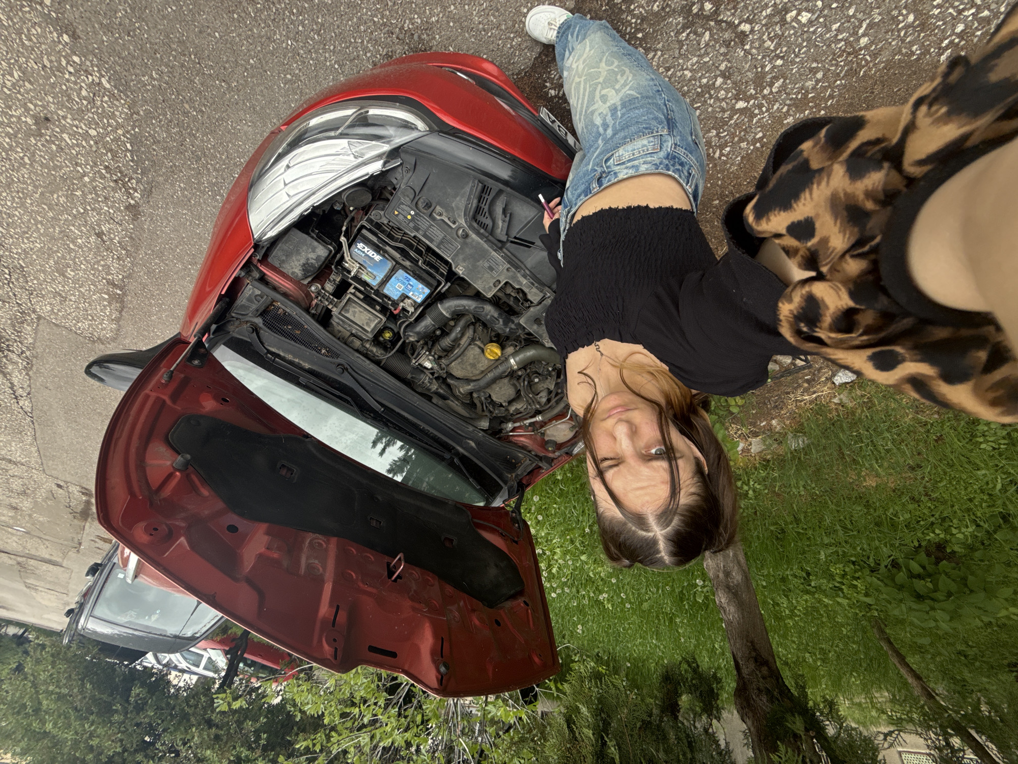 A red car with its hood open is parked on an asphalt road. A young woman stands next to the car, looking into the camera. She wears a black top, jeans, and white sneakers. Her right hand holds a pen or small tool. The car's engine compartment is visible, including the battery. A leopard-print jacket is draped over her shoulder. The background includes green grass and trees, suggesting the car is parked near a grassy area or park. Other vehicles are visible in the background behind the red car. The location is likely in Sofia, Bulgaria, based on the geotags.