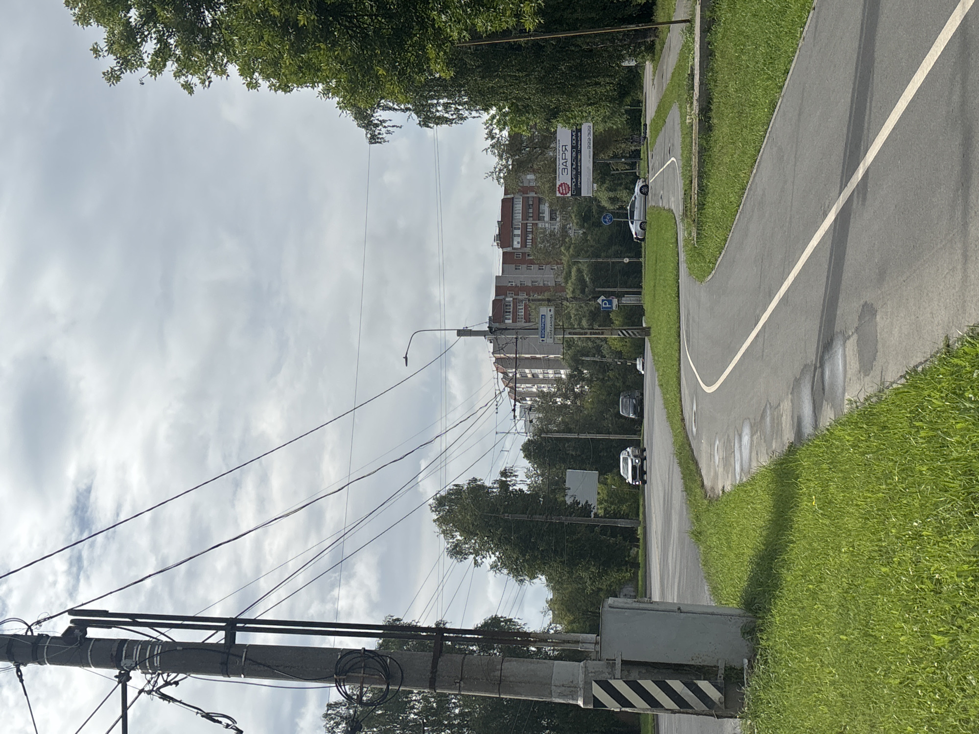 The image depicts a street scene in Ivanovo, Russia, on a cloudy day. A concrete utility pole with numerous wires dominates the left foreground. A paved road curves from the lower left towards the center and right, with a white line dividing lanes. Green grass lines the road. In the background, several cars are visible on the road, including a white sedan and a dark SUV. Behind the road, there are trees and apartment buildings. A large billboard with Russian text is also visible. The scene appears to be a typical urban intersection with pedestrian and vehicular traffic infrastructure.