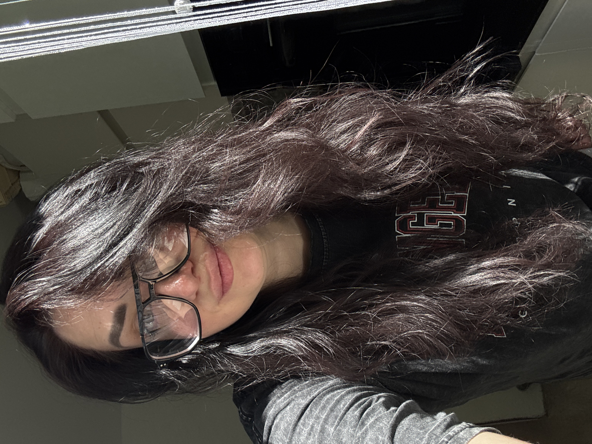A woman with long, dark hair and glasses is indoors, possibly near a window with blinds. The location is Unknown, Unknown. She is wearing a dark-colored shirt with a logo on it.