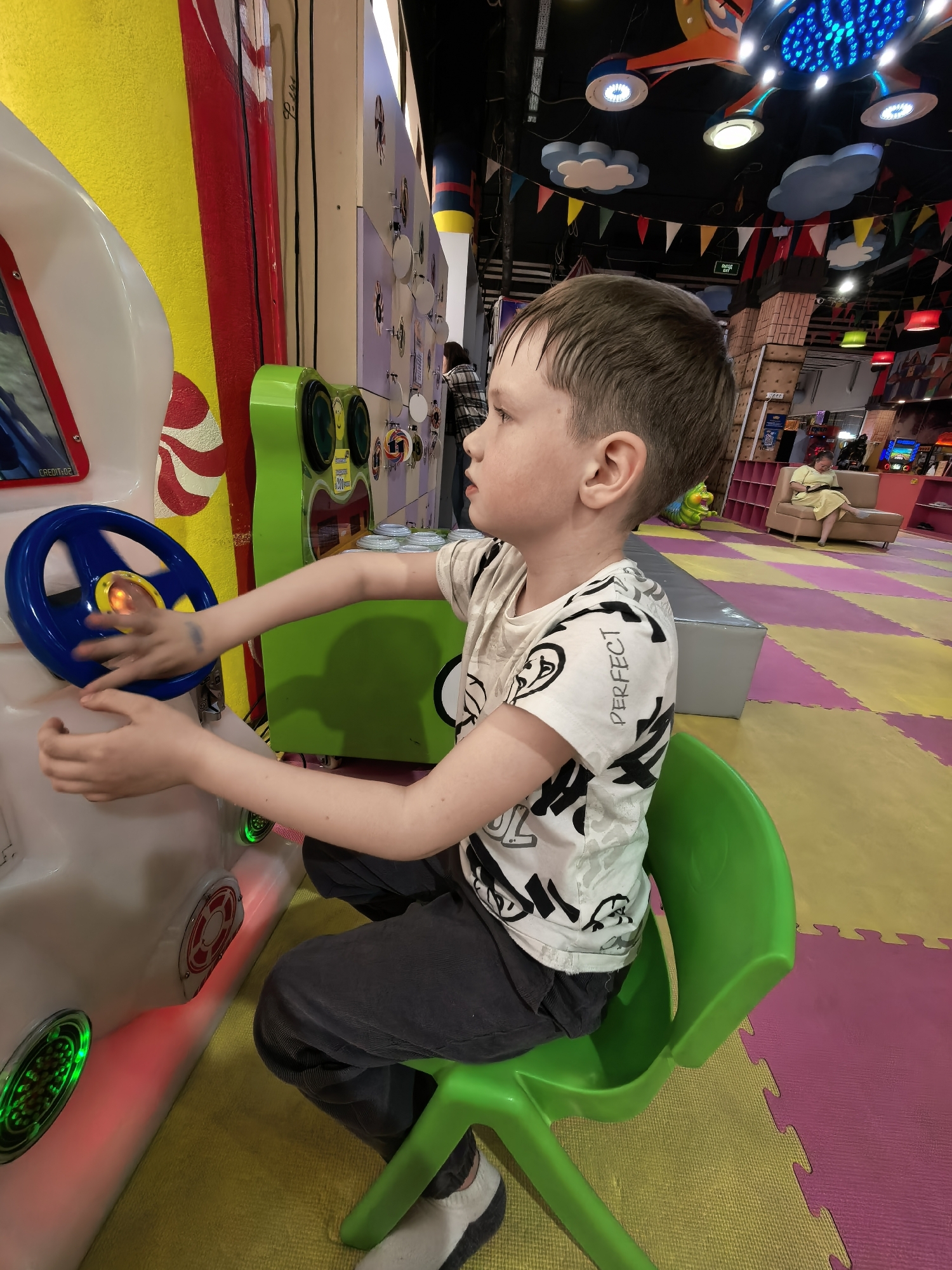 A young boy is seated in a green plastic chair in an indoor play area. He is positioned in front of a driving game, gripping the steering wheel with both hands. The game console is decorated with red, white, and yellow stripes. The boy is wearing a white T-shirt with black designs and dark pants. The floor is covered with colorful puzzle mats. In the background, there is a seating area with a woman on a sofa and various play structures under a ceiling decorated with lights and banners. The location is in the city of Unknown, Unknown.