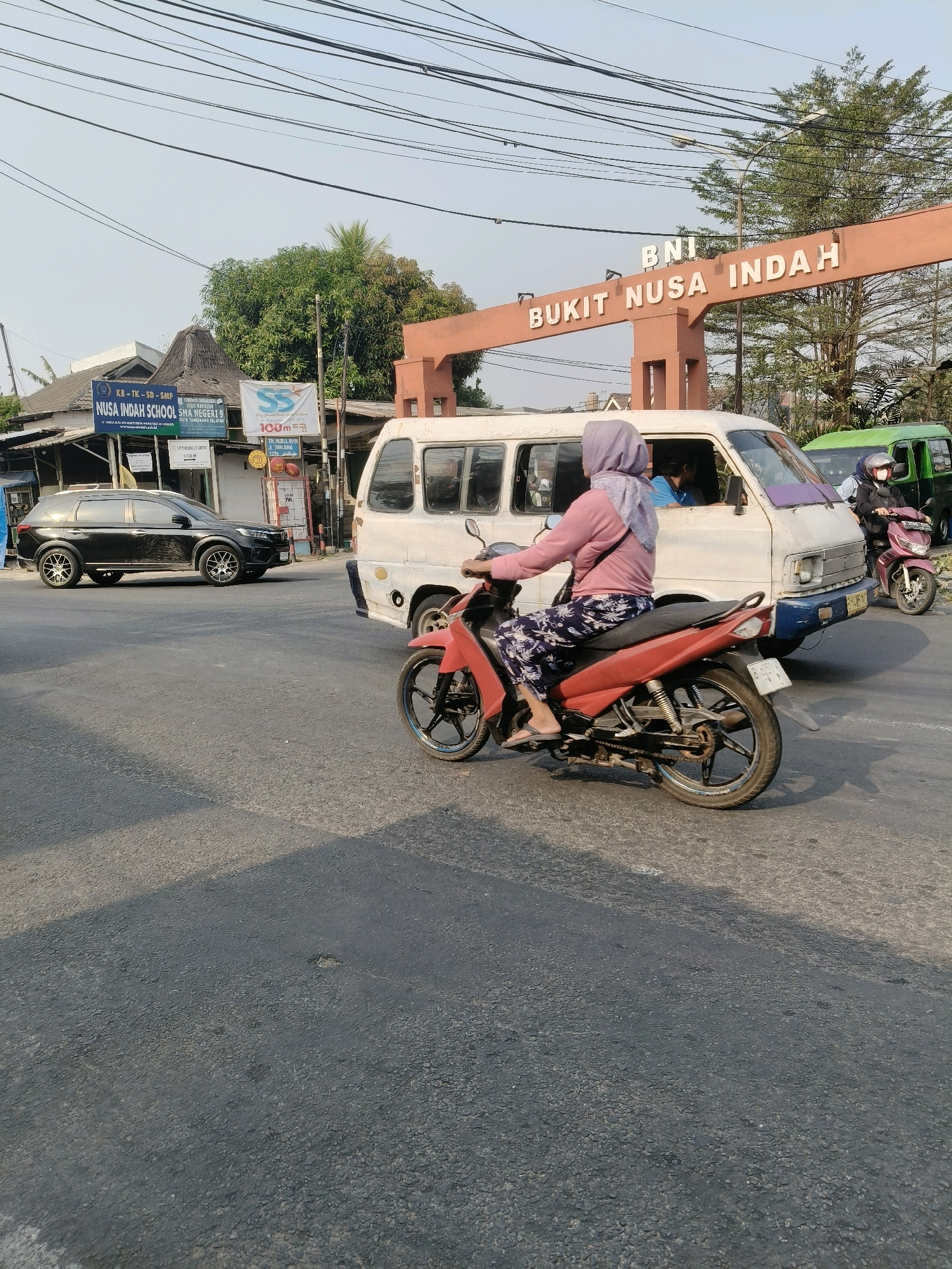 The image captures a street scene in South Tangerang, Indonesia, with a prominent archway that reads "BNI BUKIT NUSA INDAH". In the foreground, a person on a red motorcycle, wearing a pink long-sleeved shirt and patterned pants, is riding on the asphalt road. Behind them, a white van is also on the road, with other vehicles and a green truck visible further back. On the left side of the image, there are buildings with signs, including one that says "NUSA INDAH SCHOOL". Trees and utility lines are present in the background, indicating an urban or suburban environment under a clear sky.