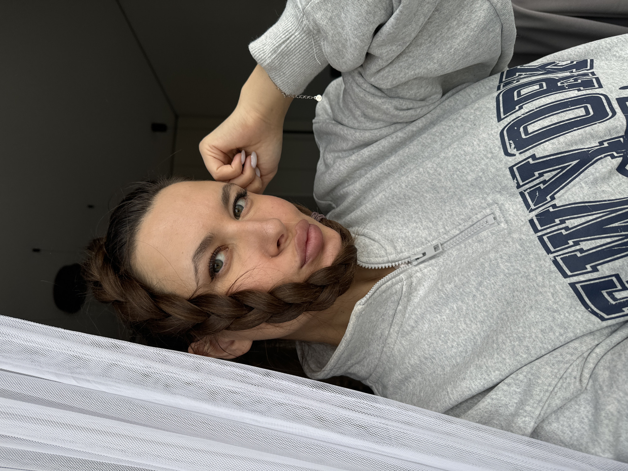 A young woman is seen from a close-up, slightly canted angle, lying on what appears to be white netting or fabric. She is wearing a grey zip-up hoodie with the word "YORK" emblazoned in navy blue lettering. Her hair is styled in a thick braid that is positioned under her chin. Her right hand is raised near her forehead, adorned with a silver bracelet. The background is blurred, suggesting an indoor setting with dark tones. There is no information available to determine if the photo was taken in Odintsovo, Russia.