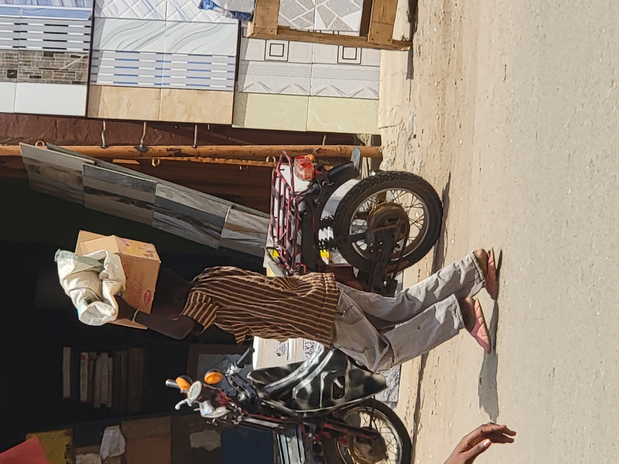 A man in a striped shirt and light gray pants is leaning over a motorcycle in Maiburiji, Nigeria. He is wearing sandals and has a box and some white cloth on the motorcycle. Behind him is a display of tiles, possibly for sale. The scene appears to be on a dusty or sandy street.