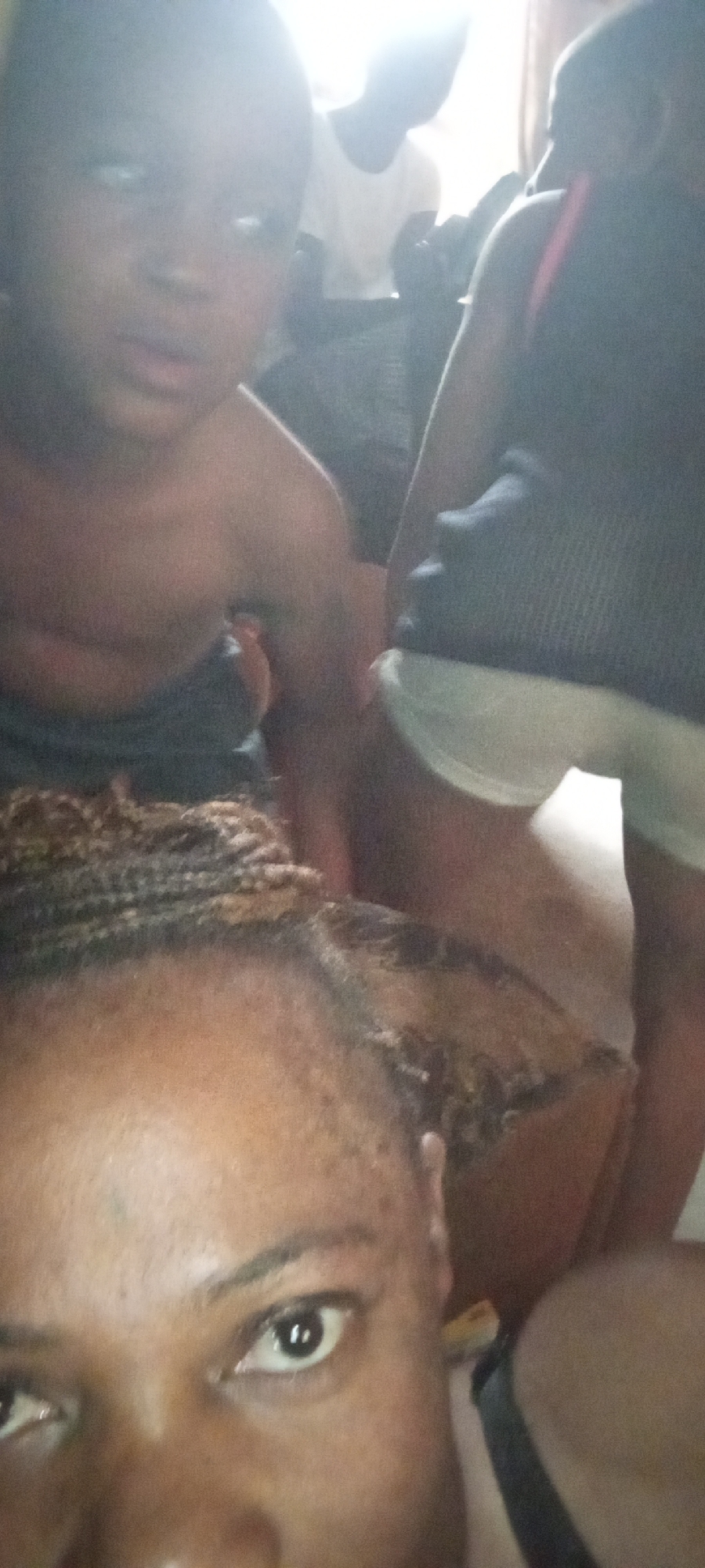 The image shows a close-up selfie of a person with dark skin and braided hair. Behind them, out of focus, are at least three other people, including one child. One person is wearing a white shirt, and another is wearing a dark tank top and light-colored shorts. The location is unknown.