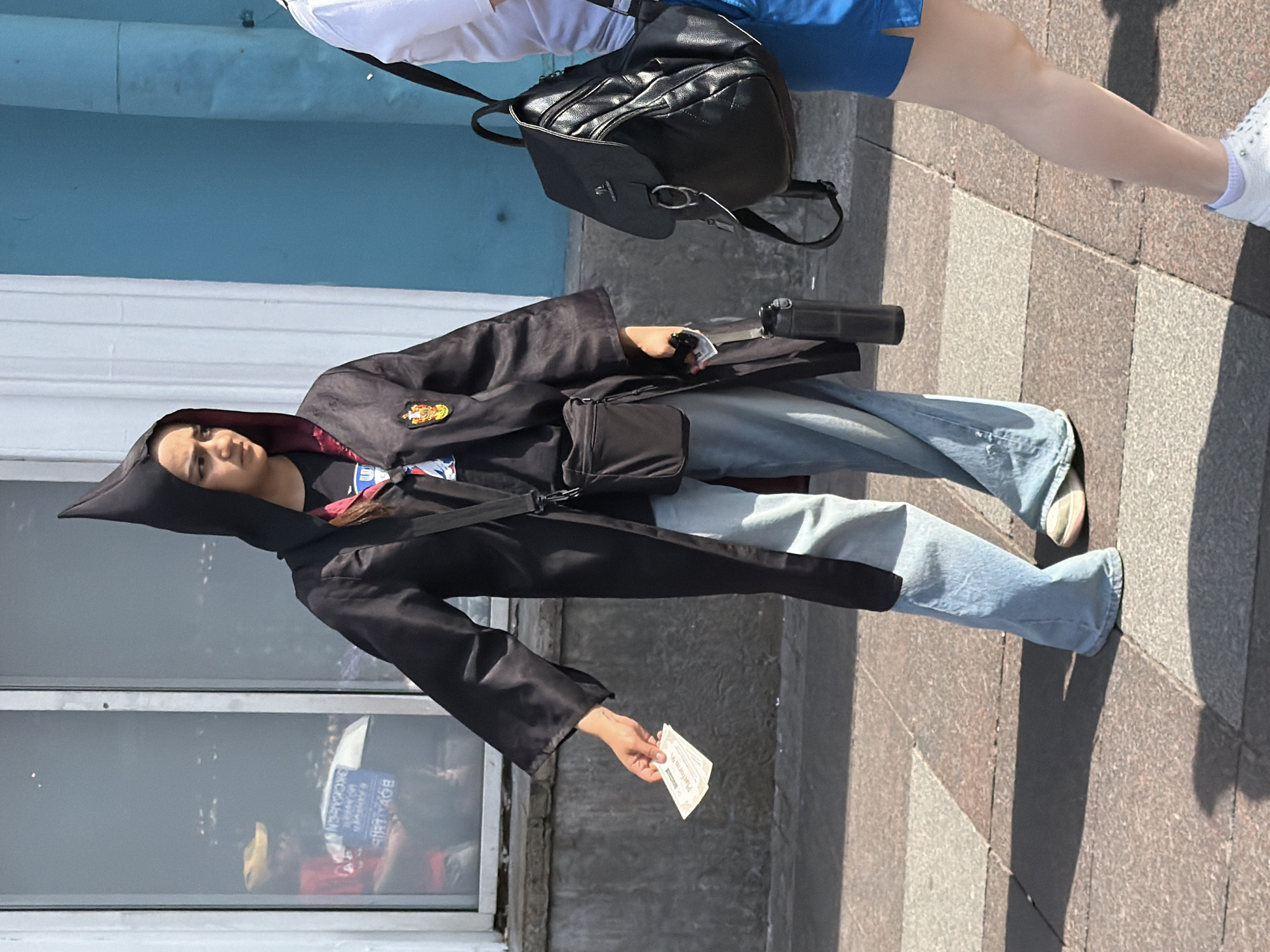 A young person dressed in a black hooded robe, reminiscent of a wizard's costume, stands on a tiled sidewalk in Saint Petersburg, Russia. They are holding two tickets in their right hand. The robe has a pointed hood and a red lining. Beneath the robe, they are wearing a black t-shirt with a crest on it and light blue, wide-leg jeans. Their footwear consists of white sneakers.
To the left of the main subject, a window reflects an indoor scene with at least two individuals. One person is wearing a yellow cap and a red shirt, holding up a blue object with text on it.
To the right of the main subject, another person, partially visible, is wearing a white t-shirt and blue shorts, with a black backpack slung over their shoulder.
A black cross-body bag is worn by the main subject, and a black water bottle is attached to their belt. The environment appears to be outdoors with architectural elements, including a blue wall and a window, suggesting an urban setting. The lighting indicates it is daytime.