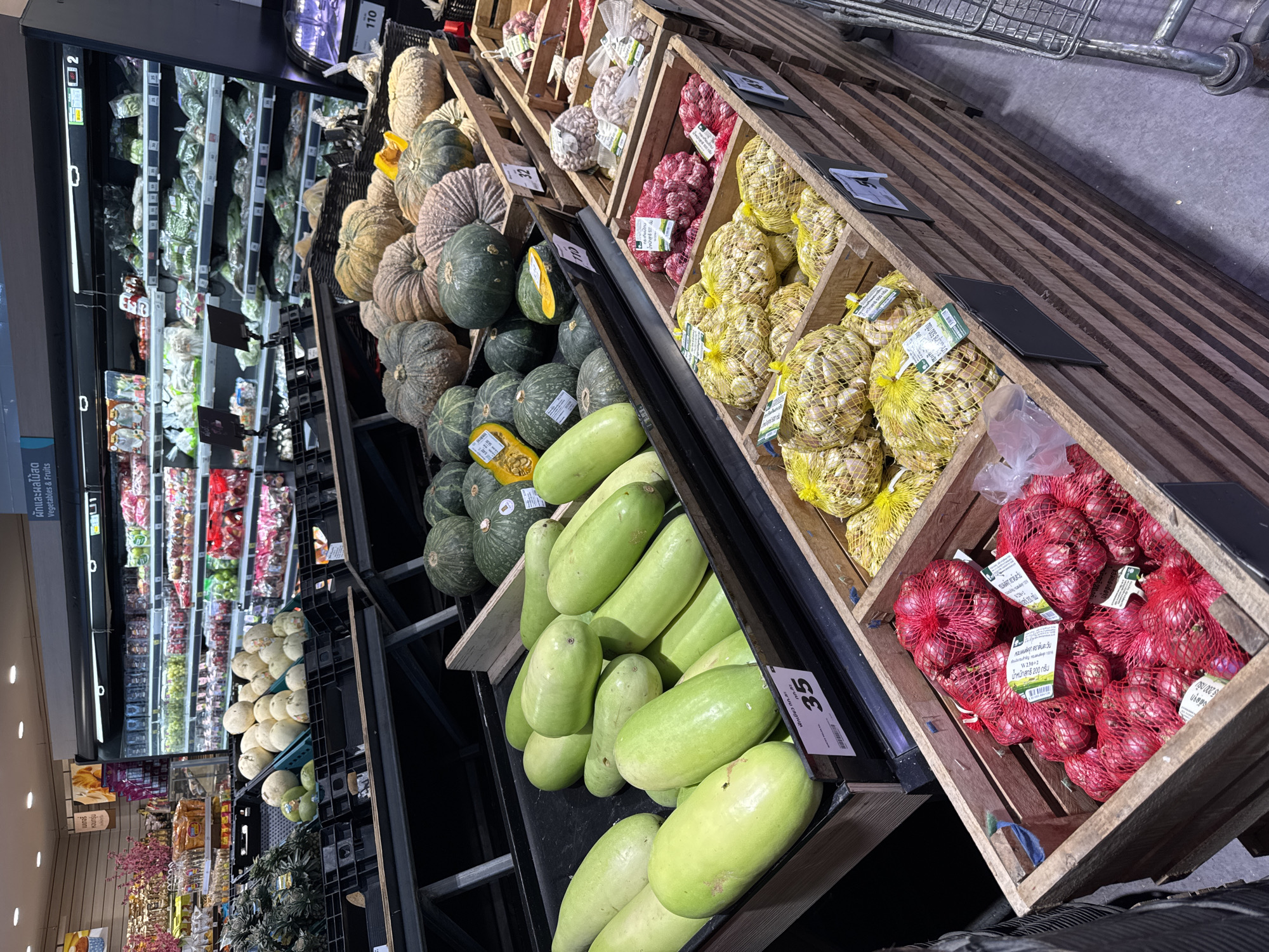 The image shows produce displayed in wooden crates in a retail environment. The produce includes pumpkins, garlic in yellow mesh bags, red onions in red mesh bags, and cylindrical gourds. Refrigerated shelving with packaged goods is visible in the background. Text on labels suggests the location is Moo 1, Thailand.