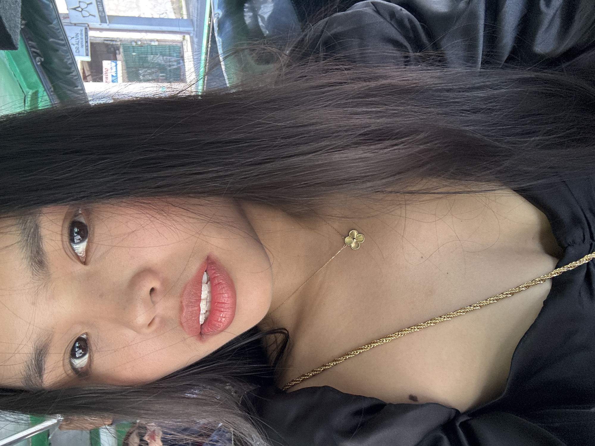 The image is a close-up, rotated portrait of a young East Asian woman, likely a selfie. She has long, straight, dark hair that cascades around her face and shoulder. Her head is tilted, and her eyes, with dark irises, are looking directly towards the camera. Her lips are parted, revealing her upper and lower front teeth, which appear to have clear dental aligners or braces. She is wearing a black, possibly silk or satin, top that drapes over her chest. Two gold-colored necklaces adorn her neck and chest: a delicate chain with a small four-leaf clover pendant rests higher on her décolletage, while a thicker, braided chain crosses diagonally across her upper chest. A small mole is visible on her upper right chest.
The background is heavily blurred but indicates an outdoor urban setting, viewed from what appears to be a shaded or covered area, possibly inside a vehicle. Hints of green trim or upholstery are visible in the foreground, suggesting this. In the distance, parts of buildings and what looks like a street are discernible under bright daylight.
Visible text on a white sign in the blurred background reads "DR. ALVIN" followed by "OPTICAL" below it, hinting at an optical shop. The overall scene suggests a casual daytime moment in a city, specifically Davao City, Philippines, given the provided context. The woman's expression is neutral, and the focus is primarily on her facial features and jewelry.