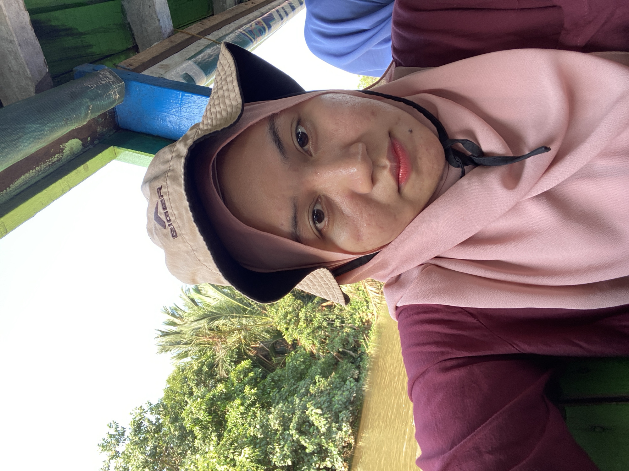 A young woman wearing a maroon shirt, a pink hijab, and a tan bucket hat is taking a selfie from a boat. She is looking directly at the camera with a neutral expression. The boat appears to be on a river in a lush, green, tropical environment, likely in Banjarmasin, Indonesia, indicated by the presence of palm trees and dense vegetation. The water is calm and appears somewhat murky. The interior of the boat is visible, with wooden beams painted green and blue overhead. Another person in a blue garment is partially visible in the background. The sky is bright and overcast.