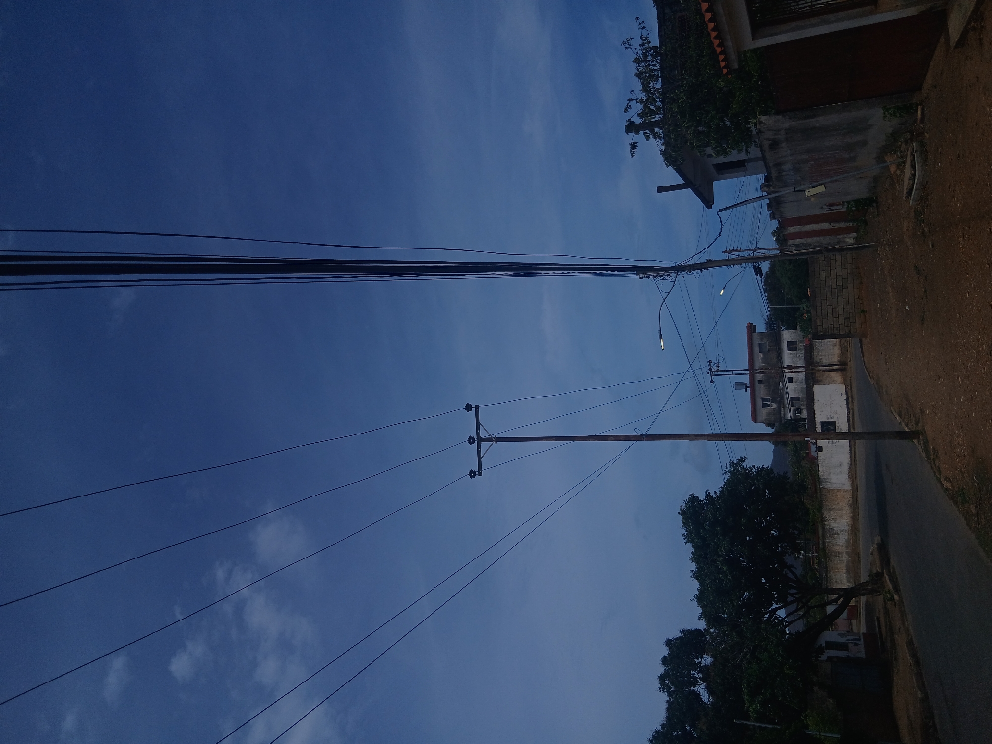 The image is taken from a low angle, looking up at a street scene in Juan Griego, Venezuela. The sky is a deep blue with some faint clouds. Overhead, there are several power lines stretching across the frame. On the right side of the image, there is a street with a few buildings visible. One building is a two-story structure with white walls and a red roof. In the lower right corner, there is a tree with dark green foliage. The overall lighting suggests it is either dusk or dawn. The streets appear to be mostly empty.