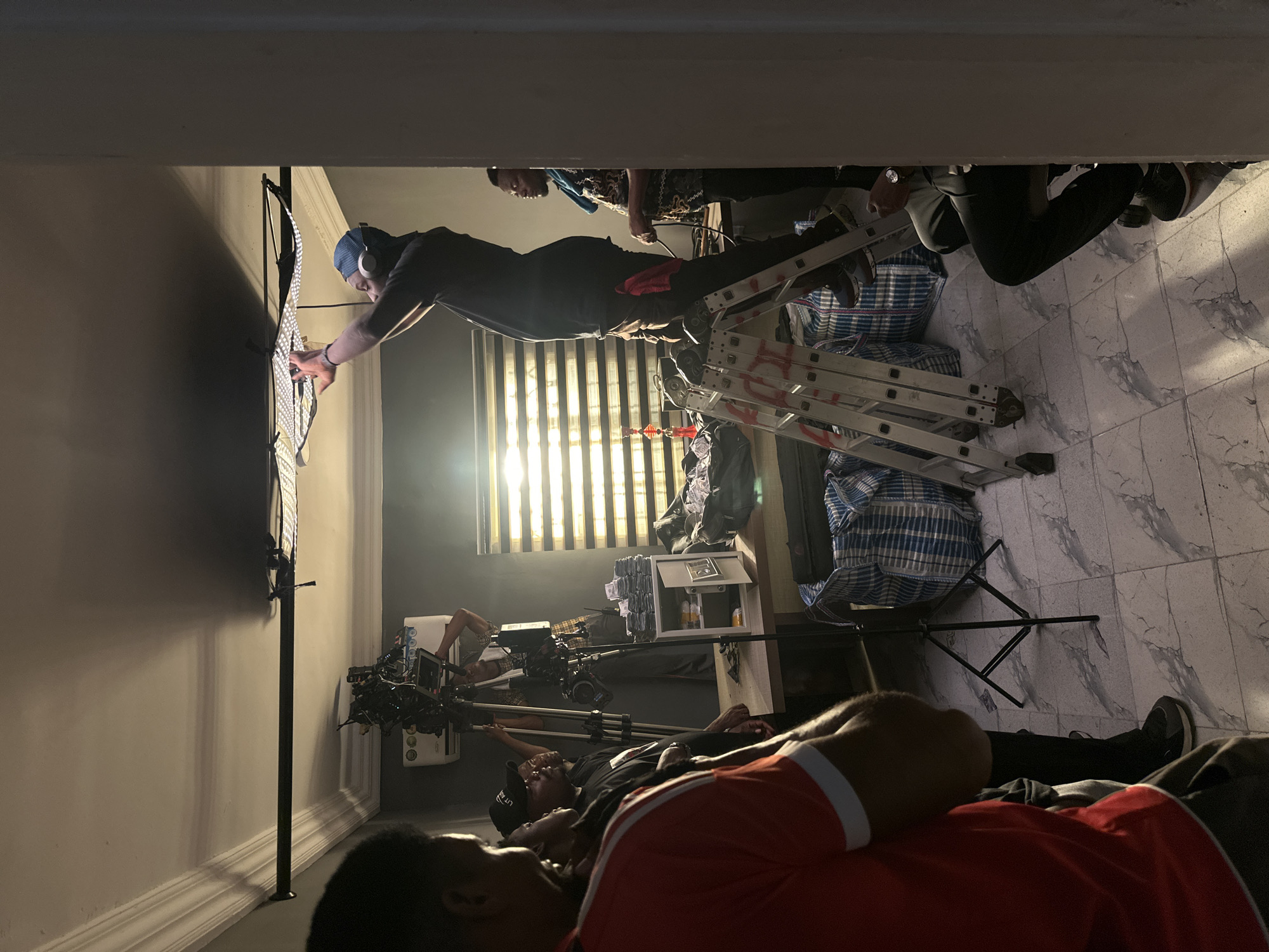 The image shows a film set in Lekki, Nigeria. A man in a blue cap and headphones stands on a silver ladder, adjusting a light panel attached to a black stand. Behind him, another man stands on the ladder. Below, a camera crew is positioned around a professional camera on a tripod. The scene is indoors with venetian blinds partially visible in the background. Bags and equipment clutter the floor. The image is rotated 90 degrees counter-clockwise.