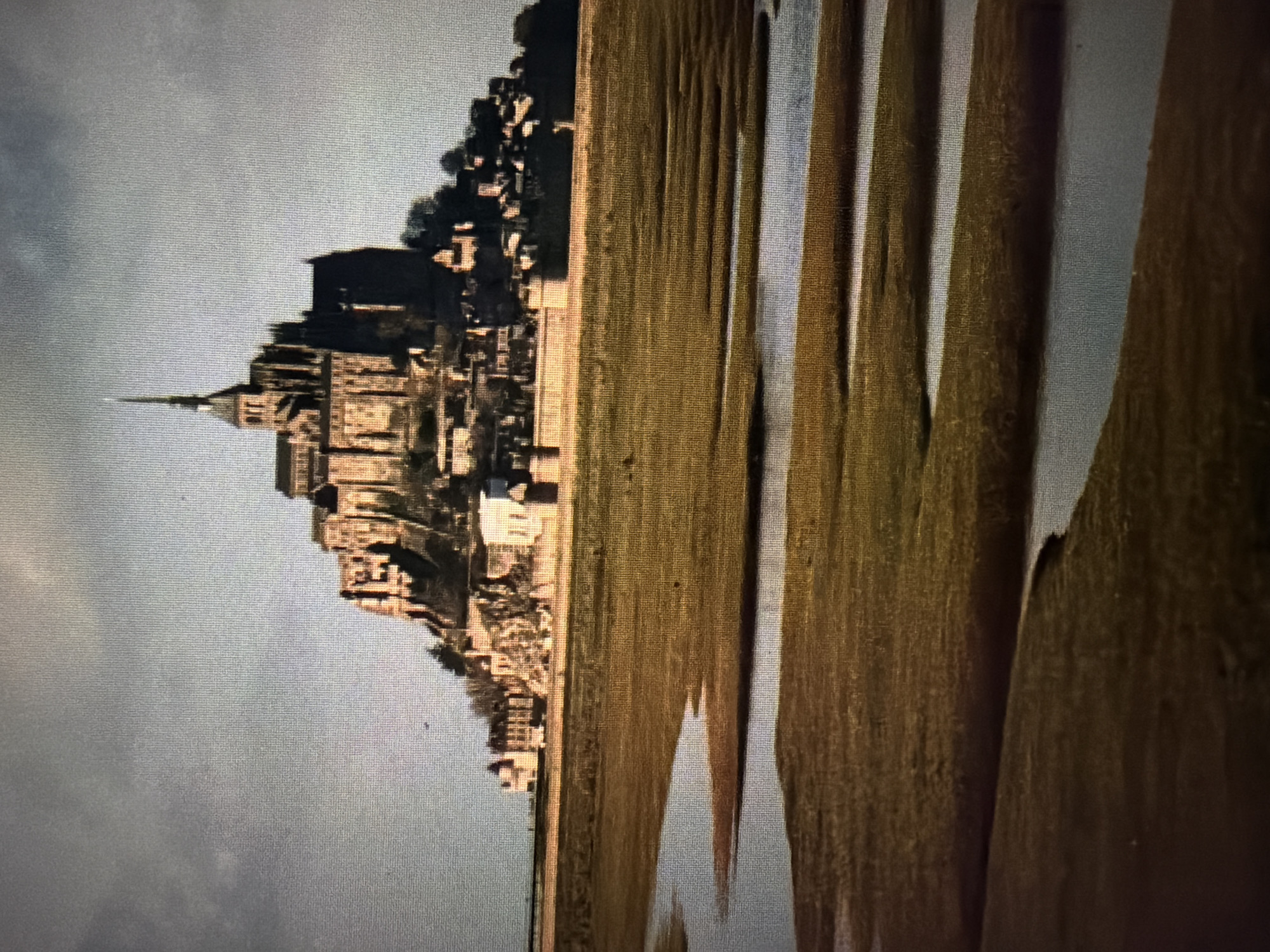 The image shows a panoramic view of Mont Saint-Michel, a medieval abbey and tidal island located in Normandy, France, not Baan Mae Nam, Thailand. The abbey is perched atop a rocky island, with its distinctive spires and architecture dominating the skyline. The foreground is occupied by a tidal marsh with winding waterways, reflecting the overcast sky. The overall scene is one of historical grandeur set against a natural, dynamic landscape.