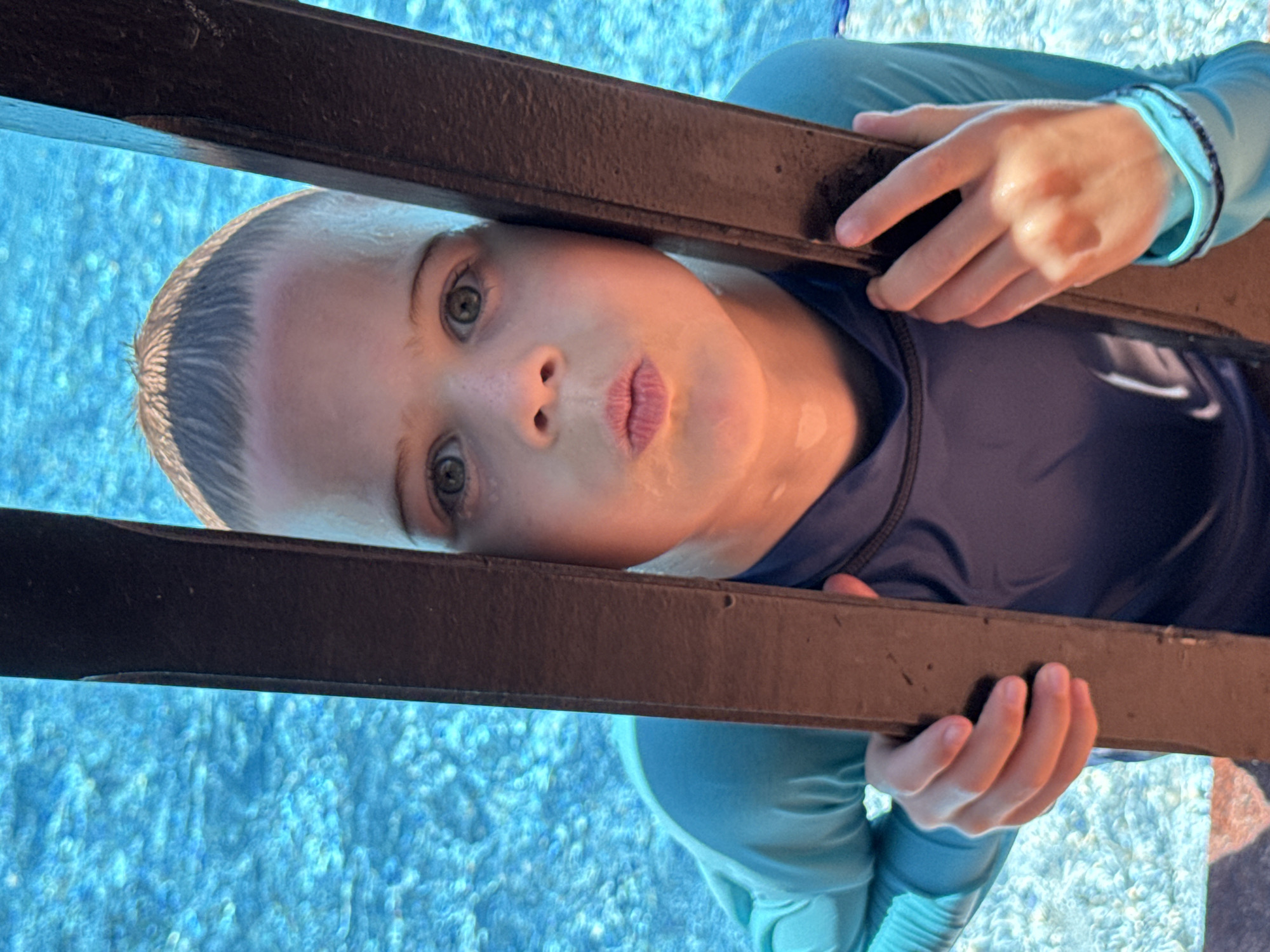 A young boy, likely Caucasian, with wet hair and a light blue rash guard over a navy blue rash guard, is looking at the camera with his lips pursed. He is standing in a swimming pool, with the blue, rippling water visible in the background. The boy is holding onto two dark brown wooden posts that are positioned vertically in front of him. His face is wet, and there appears to be some water on his skin. The lighting suggests it is daytime, and the sun is casting reflections on the water. There are no other individuals visible in the frame. The scene appears to be an outdoor swimming pool area. The location is not specified as Dubai, United Arab Emirates.