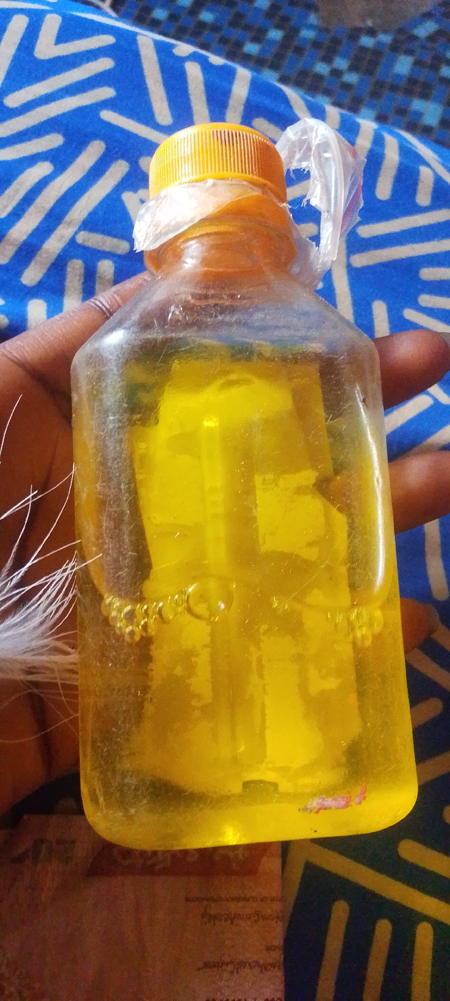 In the foreground, a person's hand with dark skin is holding a clear plastic bottle filled with a yellow liquid. The bottle has a ribbed yellow cap and a torn piece of clear plastic secured around the cap. Some small bubbles are visible in the liquid near the bottom of the bottle. White, feathery strands are partially visible to the left of the bottle. The background consists of a blue and white patterned fabric. A portion of what appears to be a pinkish-brown document with text is visible at the bottom of the image, partially obscured by the bottle. The scene is captured in a close-up shot.
