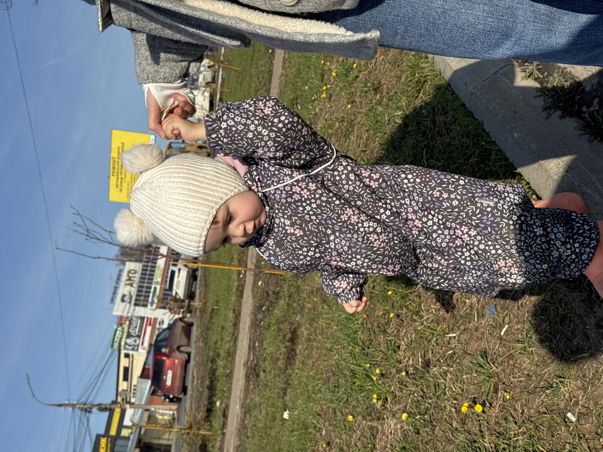 A young child, wearing a white knit hat with two pom-poms, a dark floral-patterned jumpsuit, and pink footwear, is holding the hand of an adult. The adult wears blue jeans, a grey coat, and a white undershirt. They are walking on a patch of grass with some yellow flowers growing in it, next to a concrete curb. Behind them is a city street with buildings and vehicles. The child appears to be looking down. The location is likely Chaikovsky, Russia.