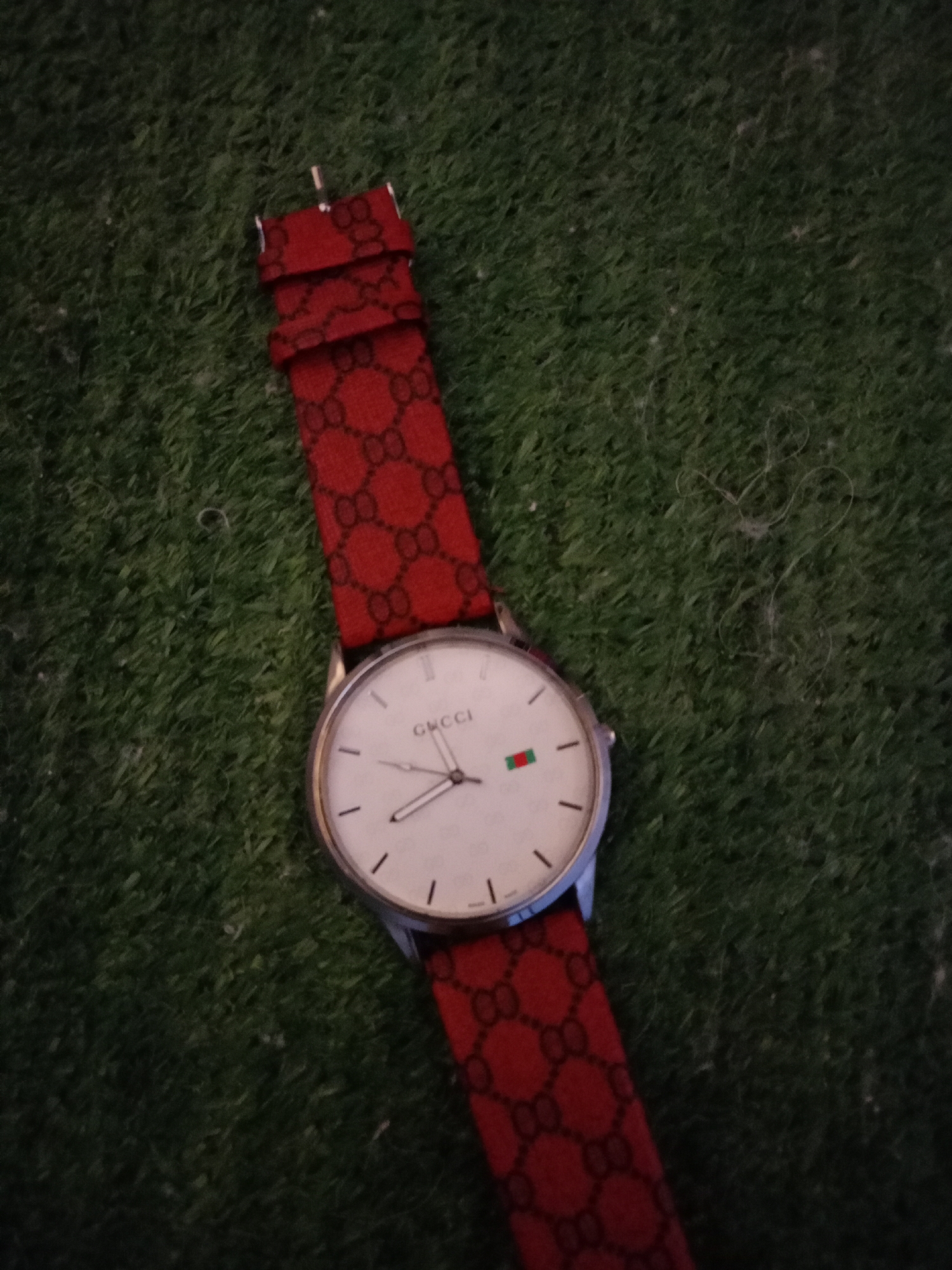 An overhead shot captures a white-faced Gucci watch with a red, patterned strap lying on a green, artificial turf surface. The watch face has thin black hour markers and two hands. A small green and red stripe is visible on the face, near the 3 o'clock position. The strap is adorned with a repeating pattern of interlocking Gs. The image is not detailed enough to determine the specific location within Petrozavodsk, Russia, or to identify any people or specific actions.