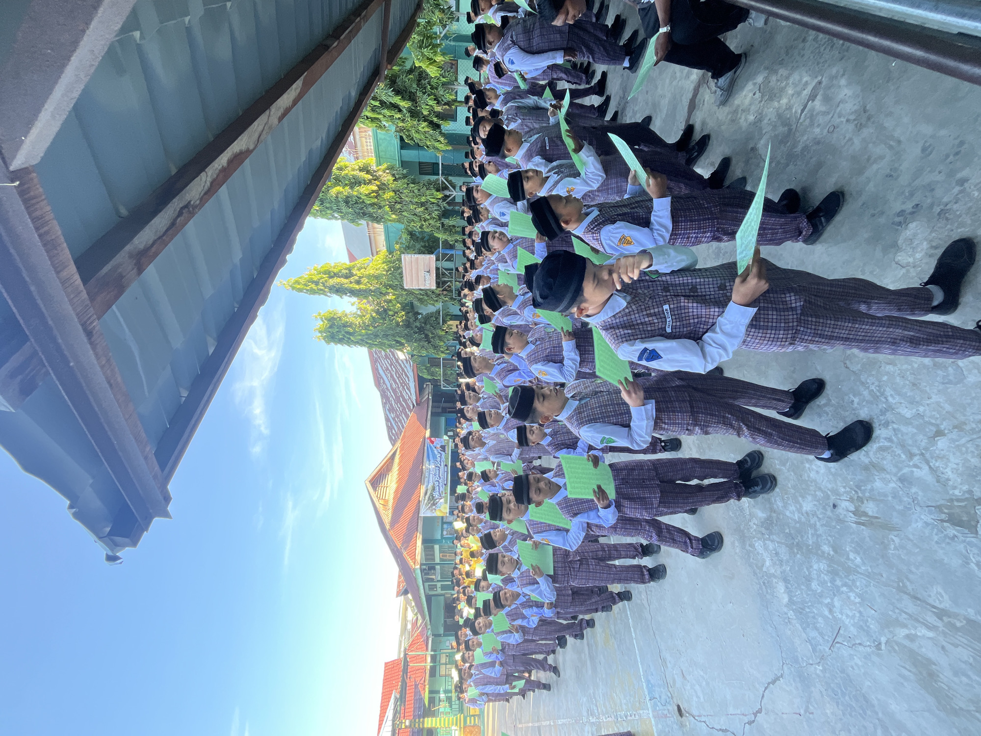 In Lhokseumawe, Indonesia, a group of young male students, dressed in school uniforms consisting of white long-sleeved shirts and plaid vests and pants, stand in neat rows outdoors. Each student holds a green piece of paper, seemingly reading or reciting from it. The students are organized in several lines, facing forward. Behind them, a building with red roofs and green walls is visible, as are tall, slender trees. The sky above is clear and blue, with a few wispy clouds. The ground is paved with concrete. The scene suggests a formal school activity, possibly a prayer, recitation, or lesson, taking place in the morning or late afternoon due to the sunlight.