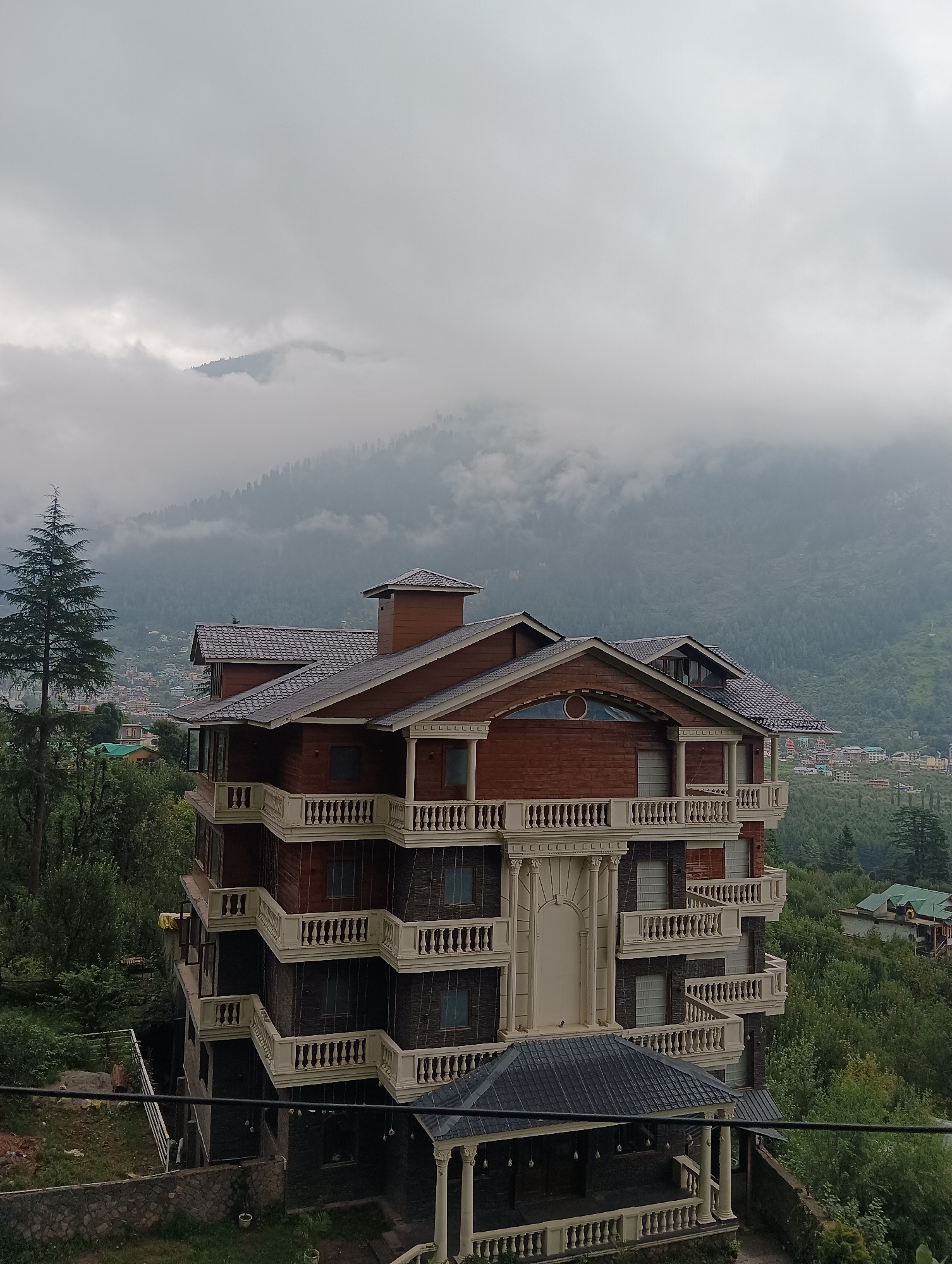 The image captures a multi-story building with a dark, tiered roof, made of wood and dark stone. The building features multiple balconies with white railings, extending outwards in a stepped pattern. A tall evergreen tree stands to the left of the building. In the background, a town is visible nestled on a mountainside, shrouded in mist. The overall atmosphere is serene and slightly overcast.