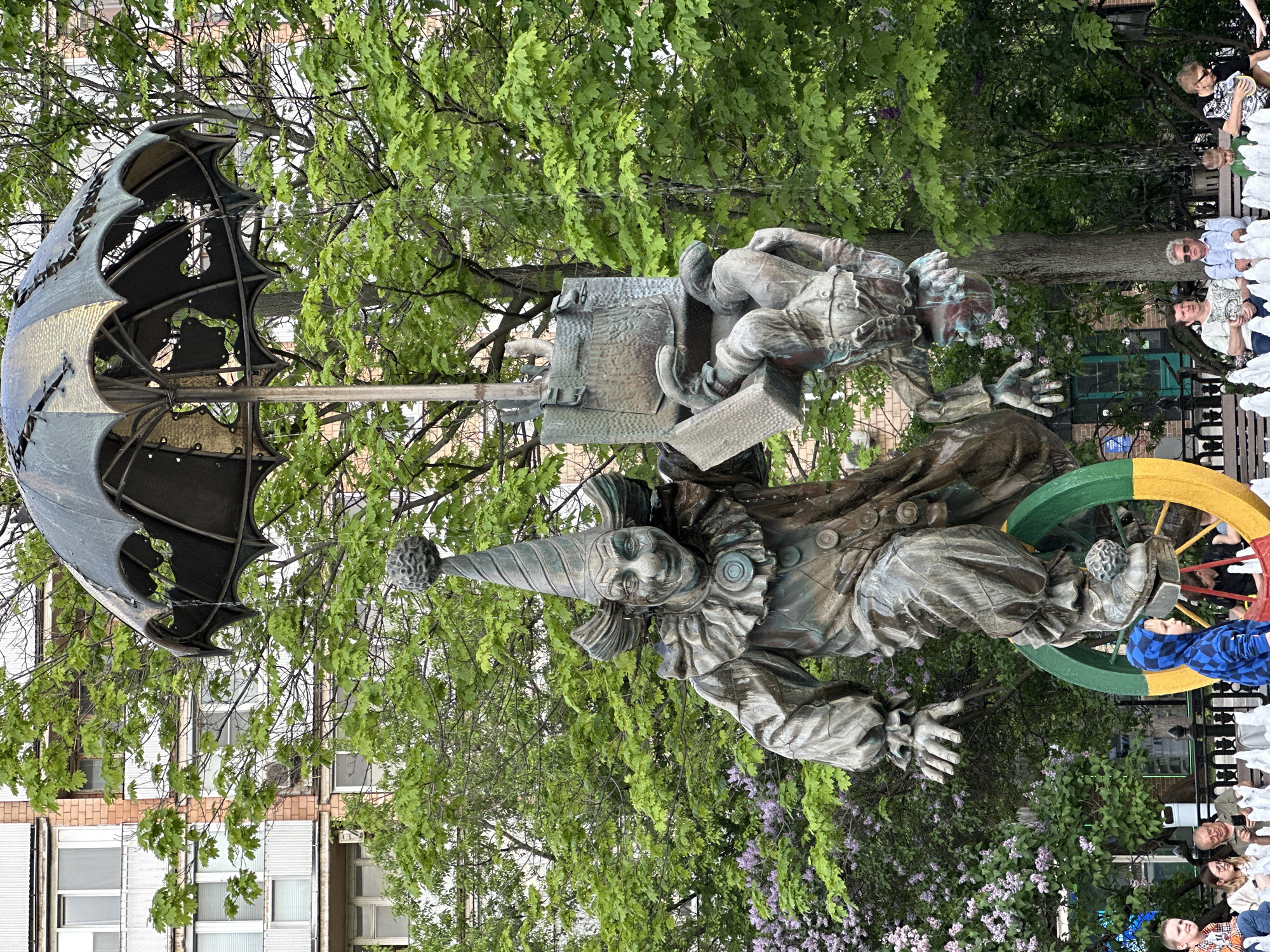 This is an outdoor scene in Moscow, Russia. The focal point is a statue of two clowns with an umbrella above them and a colorful wheel on the bottom. The sculpture appears to be made of bronze or a similar metal. The statue is surrounded by greenery, including trees and flowering bushes. In the background, there are people sitting on a bench. A multi-story building is seen in the distance.