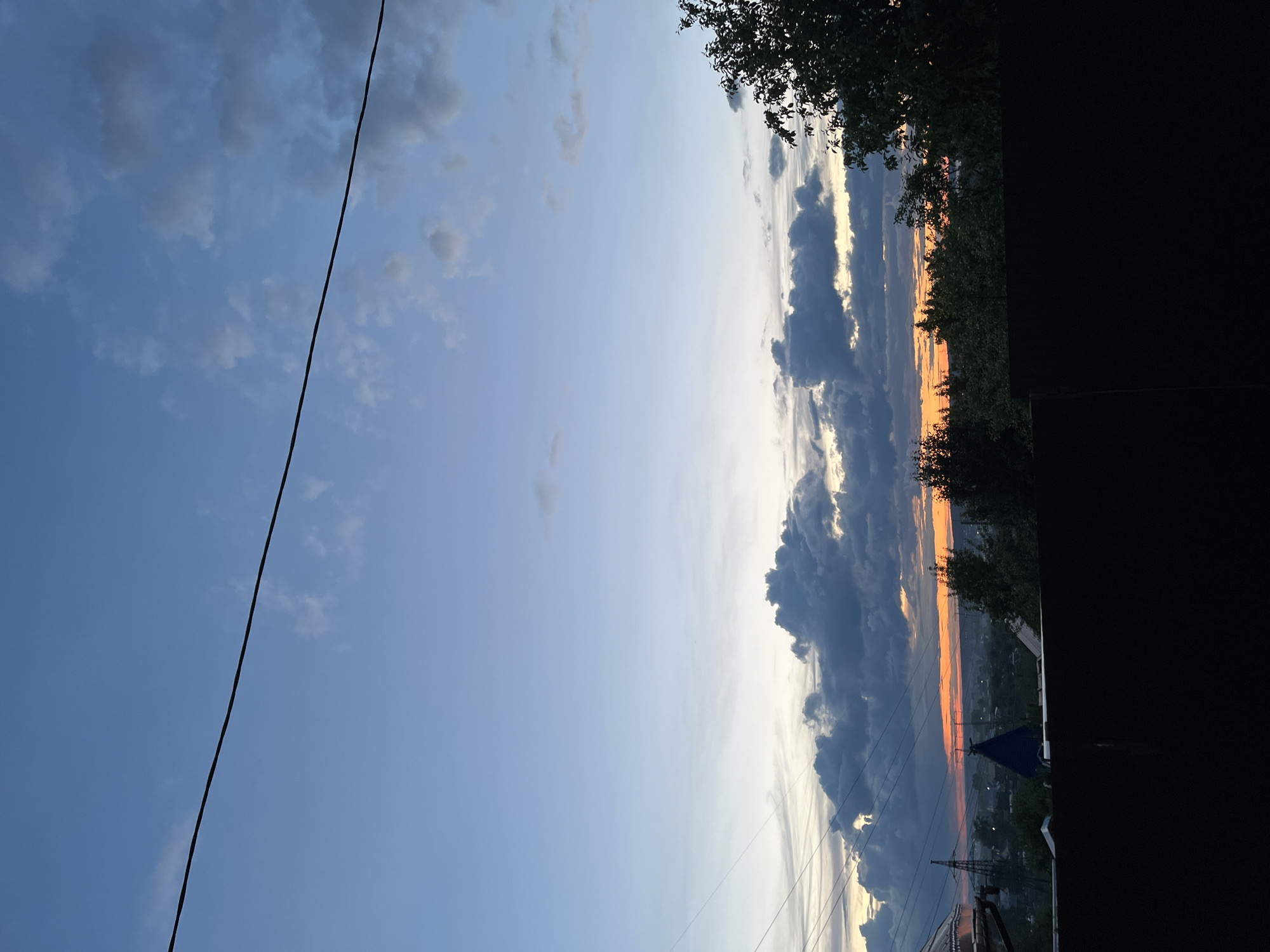 The image depicts a sky at sunset in Chaikovsky, Russia. The sky transitions from a blue hue with scattered clouds on the left to dramatic clouds with orange and yellow hues at the horizon on the right. Trees are partially visible at the top and right edge of the image, obscuring the view, along with an opaque black area covering the rightmost part. Below the sunset and clouds are some power lines, a transmission tower, and a building with a dark blue roof. A black power line traverses the upper left corner of the frame.