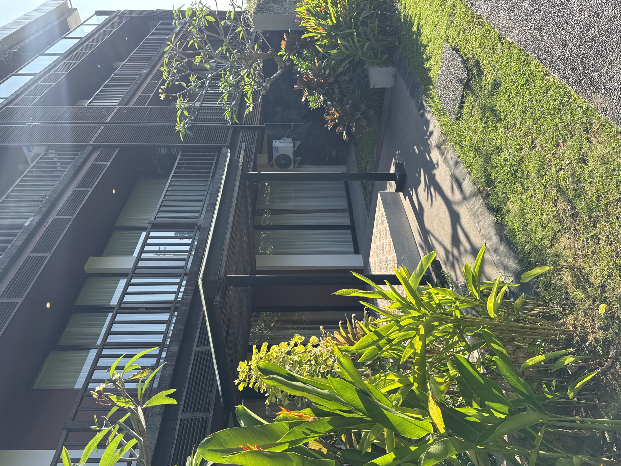 The image presents an exterior view in Kuta Selatan, Indonesia. It features a multi-story building with dark brown or black window frames and vertical slat-like structures. Large windows with white blinds or curtains are visible. Adjacent to the building is a narrow walkway made of grey concrete and gravel, alongside which are grassy areas and various tropical plants with broad green leaves. An air conditioning unit is attached to the building's exterior. The foreground contains vibrant green foliage, partially obscuring the lower portion of the building and the walkway. The lighting suggests a sunny day. No people are visible.