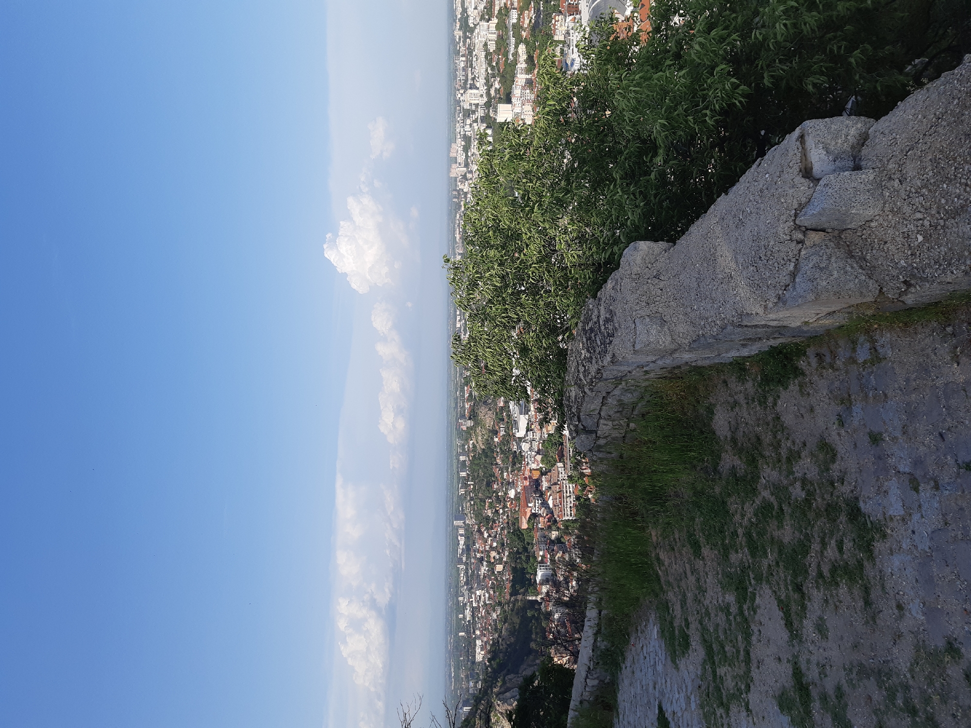 The image shows a cityscape viewed from a high vantage point, specifically from Nebet Tepe in Plovdiv, Bulgaria. The sky is bright blue with scattered white clouds. A stone wall and some green foliage dominate the foreground on the right side of the image. In the middle ground, the urban landscape of Plovdiv sprawls out, including buildings of different sizes and architectural styles. The city appears dense, and the distance to the horizon is significant.
