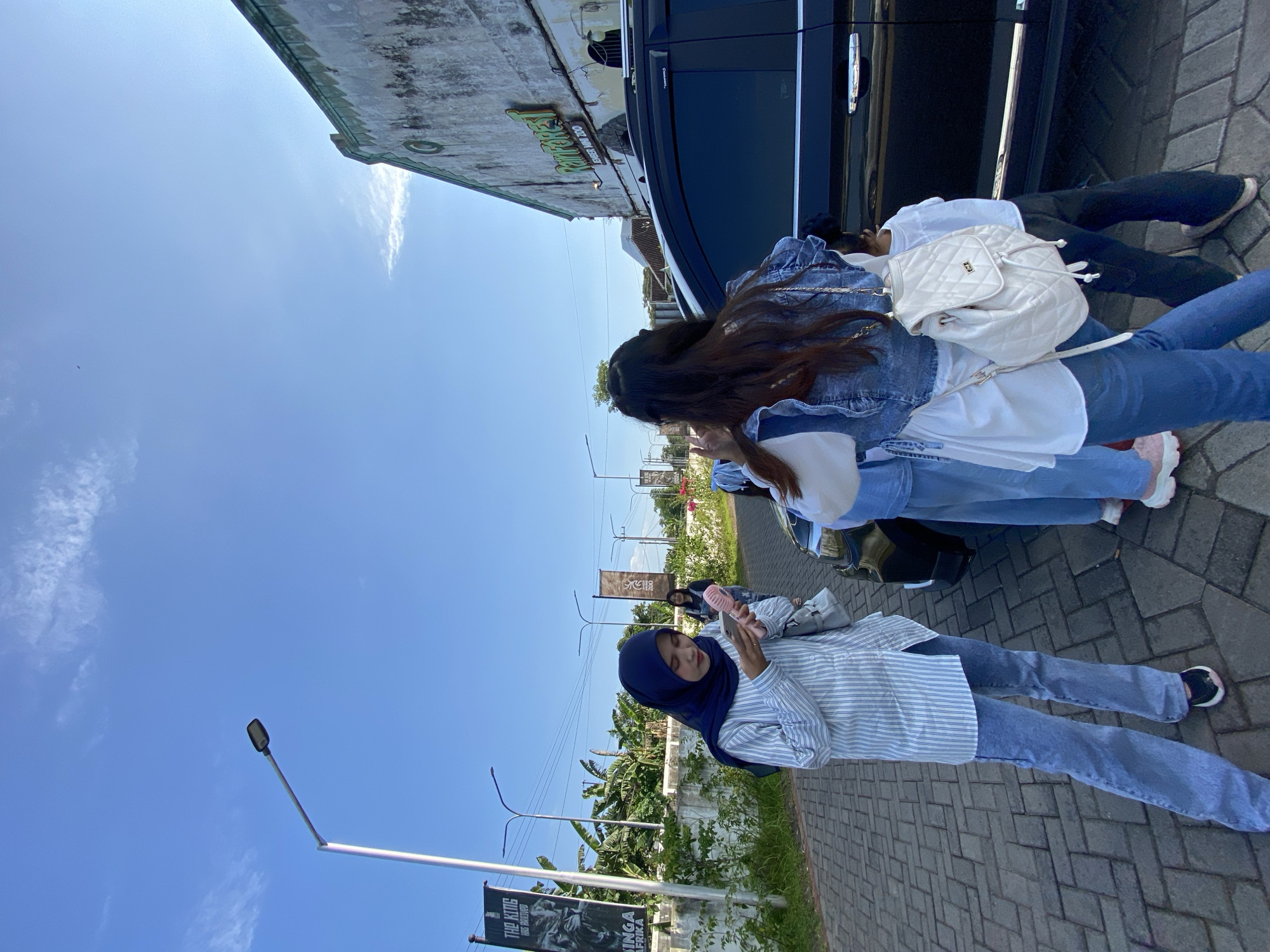 Three people stand beside a black minivan on a sidewalk paved with interlocking bricks. The person nearest the camera, a female wearing a navy blue hijab, a blue and white striped long-sleeved shirt, and blue jeans, holds a pink fan. Behind her stand two other people: one wearing a denim jacket, a white shirt, and blue jeans with a white backpack, and the other wearing a white shirt and blue jeans. The background includes a blue sky, a building with signage in a language other than English, street lights, banners, and foliage. This photo was taken in Mangli, Indonesia.
