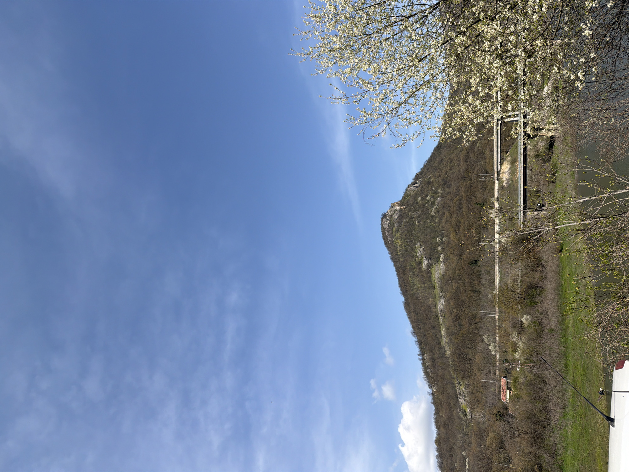 The image shows a landscape in Asparuhovo, Bulgaria. A mountain covered in dry vegetation rises vertically from the right side of the image. On the side of the mountain, there is a narrow road or path. A white pedestrian bridge spans a body of water adjacent to the mountain. In the upper right corner of the image, there are branches with white blossoms, indicating springtime. The sky above the mountain is a clear blue with some scattered wispy clouds. At the bottom right, a white object and a black antenna are visible.
