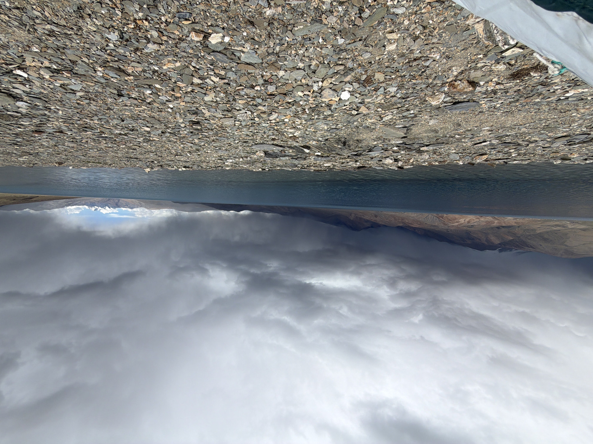 A wide-angle, eye-level, outdoor, daytime, horizontal shot captures a desolate, rocky shoreline with a vast, dark blue lake stretching towards a distant mountain range under a dramatic, overcast sky. The foreground is dominated by a dense carpet of various-sized, flat, grey and brown stones, interspersed with a few white and reddish rocks. The shore slopes gently towards the water. The lake's surface shows gentle ripples, and its color deepens to a darker shade as it approaches the distant mountains, which are rendered in muted browns and greys. Directly above the mountains, the sky is filled with heavy, dark grey clouds, some of which appear to be letting loose rain, creating dark vertical streaks that blend with the mountain shadows. Lighter grey clouds are visible higher up, and in the far right, a small patch of lighter sky hints at a possible break in the storm. A white, tarp-like object, possibly part of a shelter or vehicle, is visible in the lower left corner, partially obscuring the shoreline. The scene conveys a sense of rugged, wild, and somewhat ominous natural beauty, possibly in a high-altitude region. The location is identified as Pangong Tso, Ladakh, India.