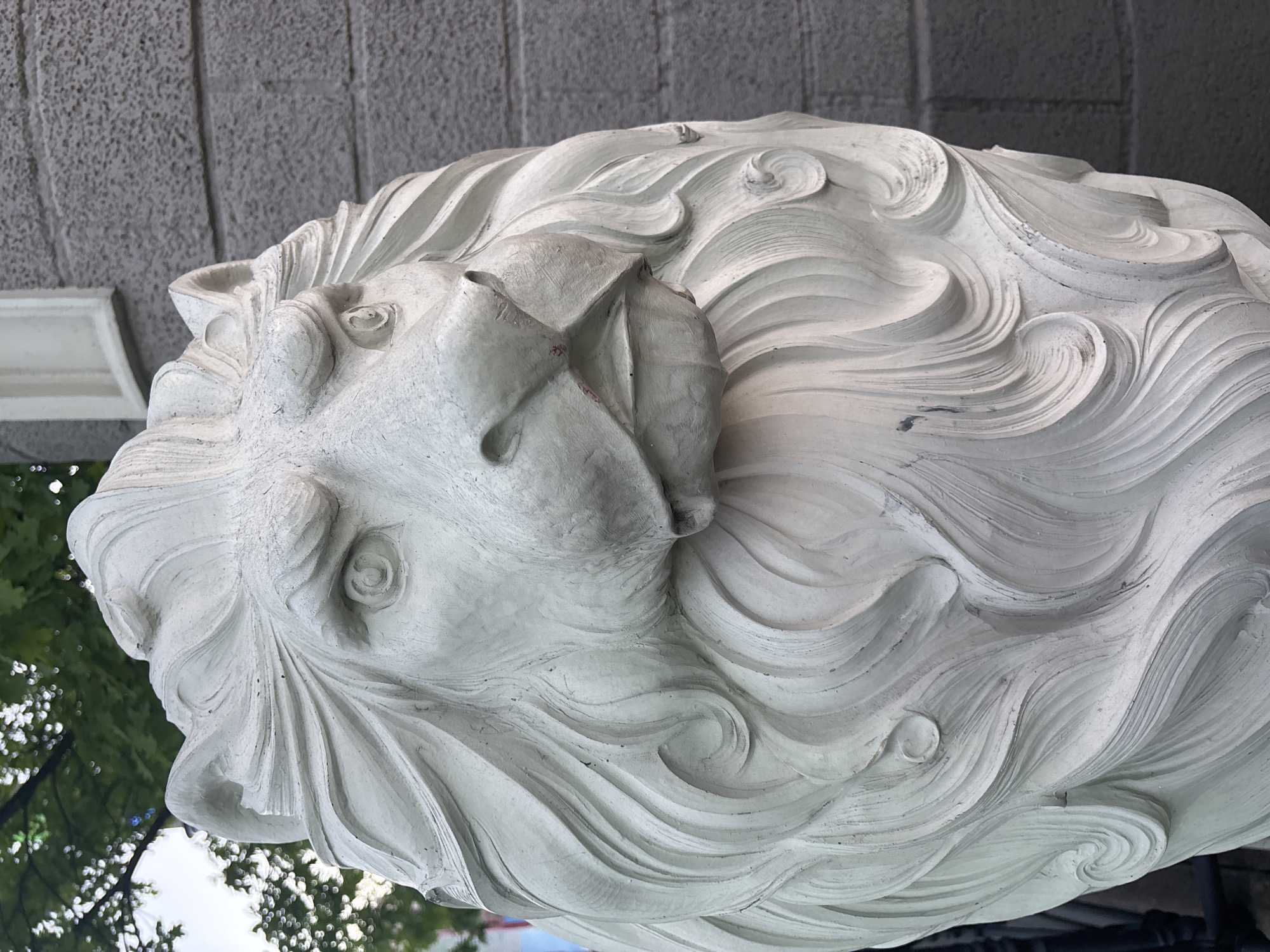 The image shows a close-up of a white, sculpted lion head. The statue appears to be made of stone or plaster, with detailed carvings of the lion's mane. The lion's facial features are visible, including the eyes, nose, and mouth. The background includes a grey concrete or stone wall and some greenery, suggesting the statue is located outdoors. The location is Moscow, Russia.