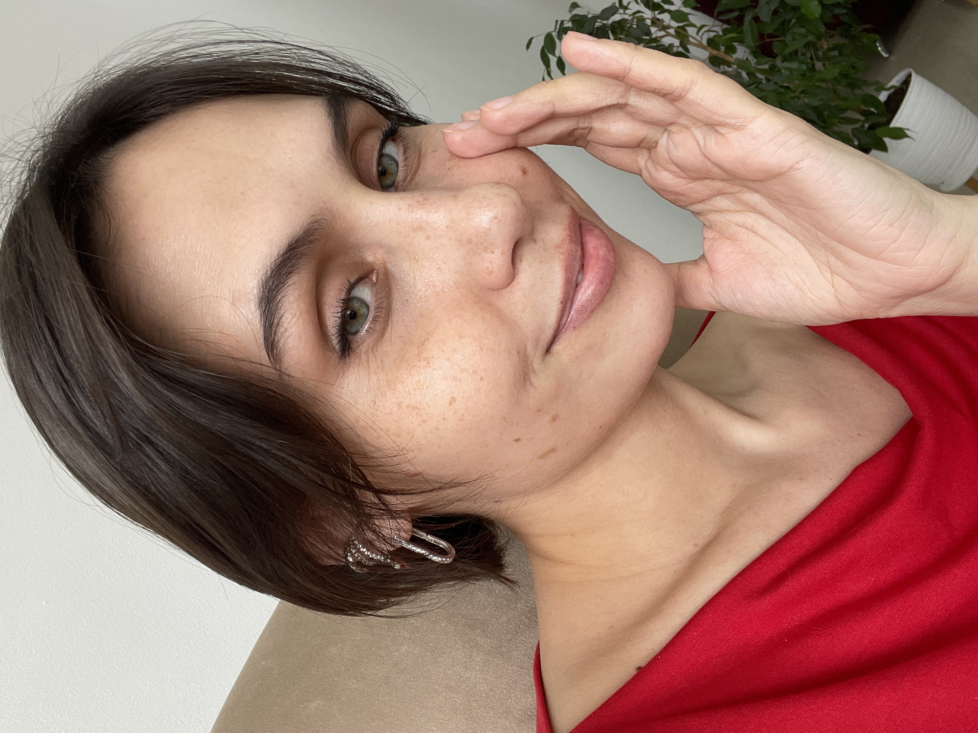 A close-up portrait of a young woman from Izhevsk, Russia, looking directly at the viewer. She has short, dark brown hair, dark eyebrows, and striking green eyes. Her fair skin is notable for the scattering of freckles across her nose and cheeks. She is wearing a vibrant red top or dress, with a glimpse of her bare shoulder. A silver-toned, possibly ornate, hoop earring is visible in her left ear. Her left hand is gently raised to her face, with her fingers softly touching her temple and cheek. Her expression is calm and natural, conveying a serene or contemplative mood. The immediate setting suggests she is leaning on a light brown or beige upholstered surface. The background features a plain white wall, with a potted plant, likely a Ficus, in a white pot visible on the right side of the frame. The lighting appears soft and diffused, indicating an indoor shot during daytime. No text is visible in the image.