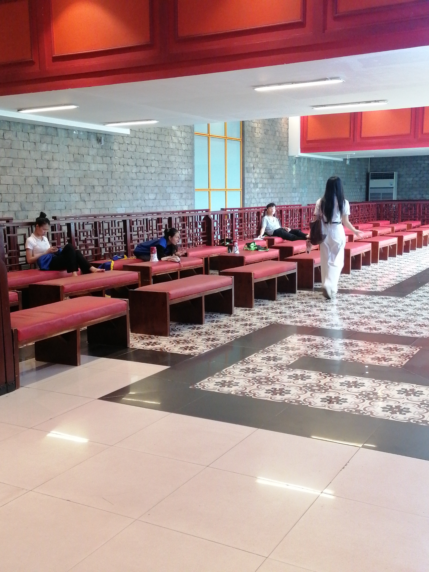 The image depicts an indoor setting with several people, likely in a public space within Bai Chay Ward, Vietnam. The foreground features a tiled floor with a pattern of light-colored squares interspersed with sections of decorative floral designs in dark tones. Arranged along a low, ornate railing are a series of low, red cushioned benches. Three women are seated or reclining on the benches, engaged in various activities such as using their phones. A fourth woman, seen from the back, is walking away from the camera towards the back of the room. The room is capped by a red horizontal bar/frame at the top of the wall. The architecture includes a brick wall in the background and a window with gold trim is also present. The general environment appears to be a waiting area or lounge.