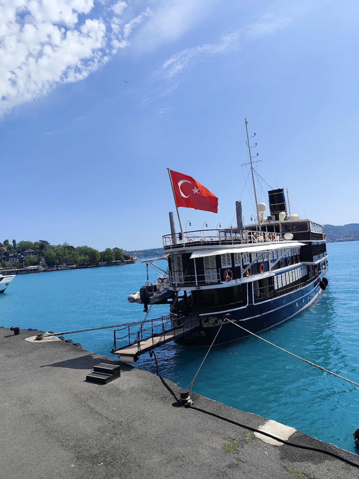 The photograph captures a maritime scene in Sarıyer, Turkey, showcasing a large, multi-decked yacht docked at a concrete pier. The vessel, predominantly black with white accents and numerous life preservers visible along the sides, is adorned with the Turkish flag fluttering from its stern. A smaller dinghy is attached to the rear of the yacht. A gangplank extends from the yacht to the pier, facilitating access. The pier is constructed of dark gray concrete and features a set of steps leading up to it. Ropes secure the vessel to bollards on the pier. The water surrounding the yacht is a vibrant turquoise. In the background, a verdant shoreline with buildings nestled among the trees is visible under a clear blue sky with scattered white clouds.